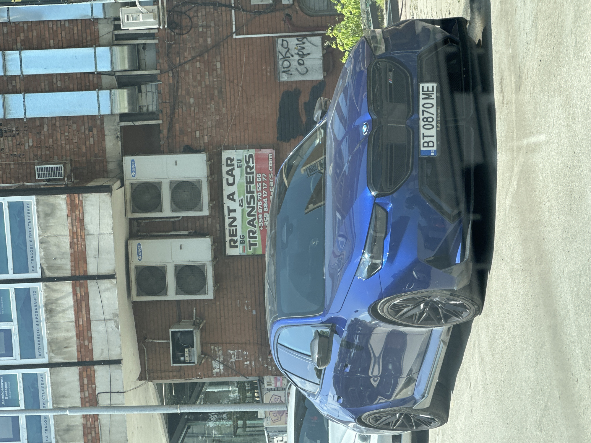 Here is a description of the image:
The image shows a blue BMW car parked in front of a brick building in Veliko Tarnovo, Bulgaria. The license plate number of the car is BT 0870 ME. On the building, there are several air conditioning units, as well as signs for "RENT A CAR & TRANSFERS" and political advertisements for the "Democratic Bulgaria" coalition. There is also graffiti on the building.