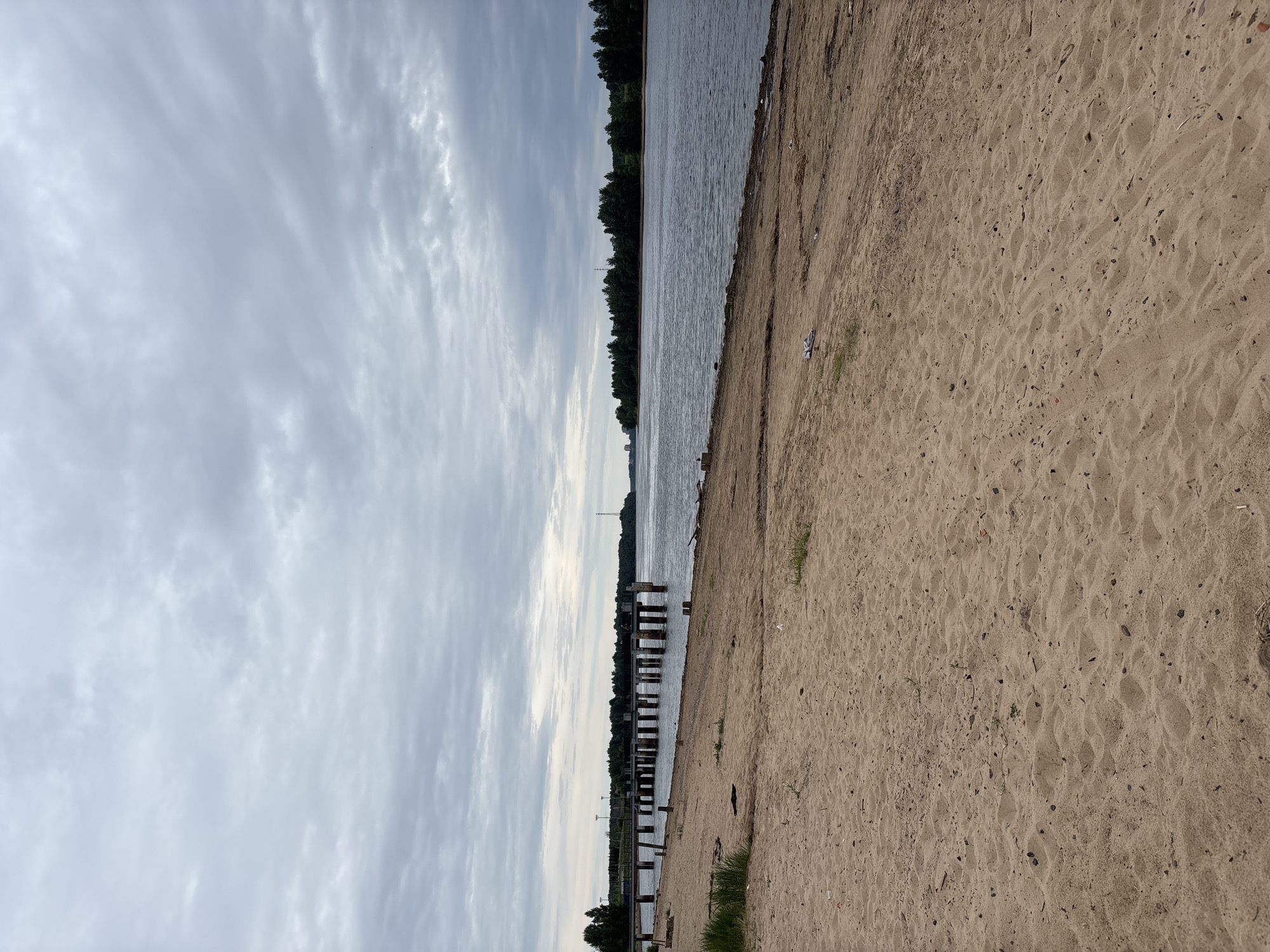 The image shows a sandy beach in Nevskaya Dubrovka, Russia, with a wide body of water in the background. A dilapidated pier, with wooden pilings and concrete structures, extends out into the water from the left side of the frame. Trees line the opposite shore, and a transmission tower is visible in the distance. The sky is overcast with gray clouds. The sand in the foreground is textured with ripples and scattered debris, including what appears to be a pair of white shoes and some plastic. The water is calm and reflects the cloudy sky. The overall scene suggests a somewhat neglected, natural waterfront environment. There are no people visible in the image.