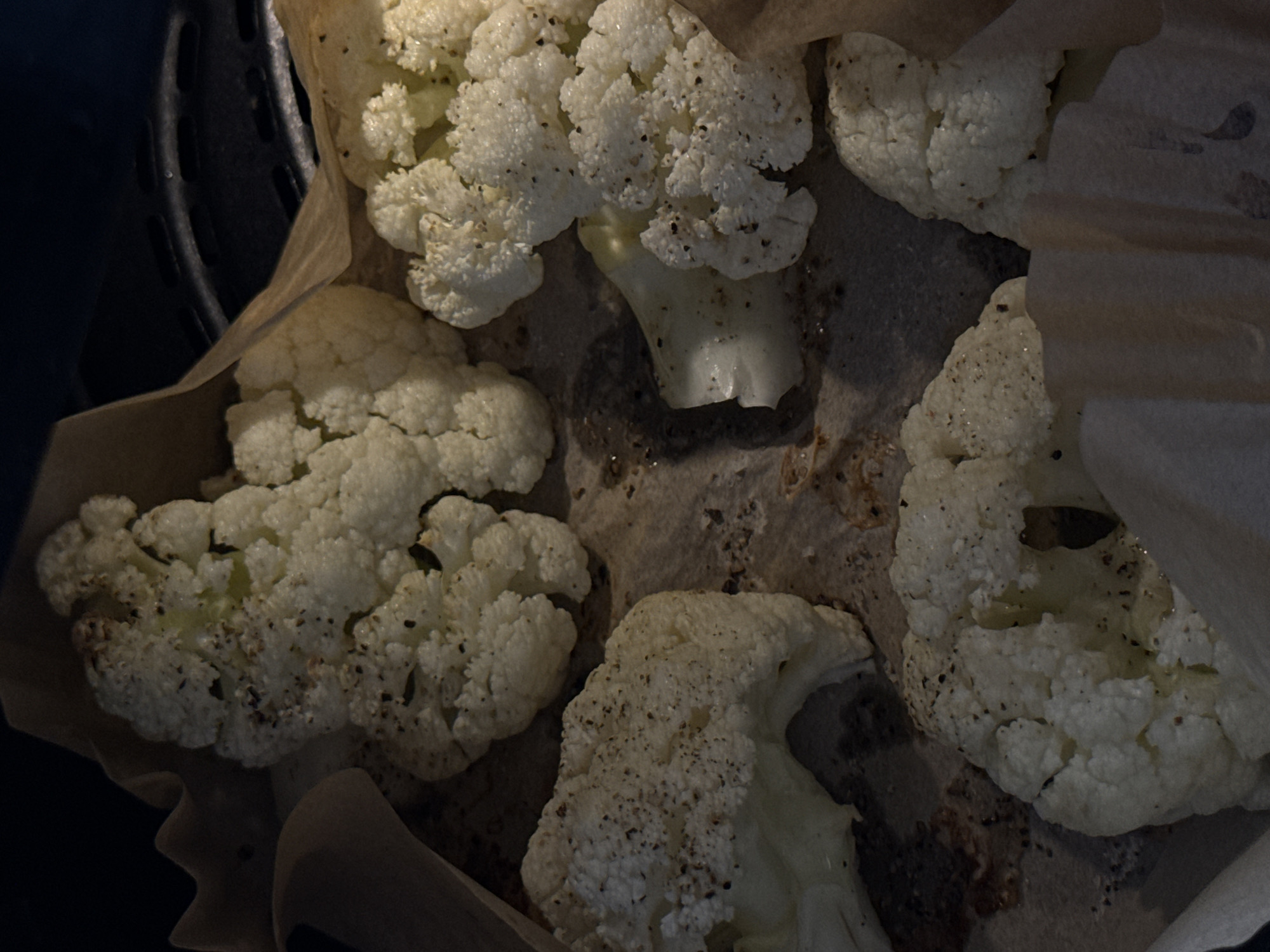 This close-up image shows several white cauliflower florets, seasoned with dark specks that appear to be ground black pepper, positioned inside what looks like an air fryer basket. The cauliflower rests on a crumpled, light brown parchment paper liner, which conforms to the shape of the cooking appliance. A dark, ribbed edge of the appliance's basket is visible on the left side of the frame, indicating an air fryer or similar cooking device.
The florets vary slightly in size and shape, with their characteristic bumpy, textural surface clearly visible. Some of the florets show hints of their light green stems. The background beneath the parchment paper appears dark and somewhat stained, consistent with a cooking environment. The lighting is somewhat dim, suggesting an indoor setting under artificial light, but it is sufficient to highlight the details and texture of the cauliflower and seasonings. No people, visible text, specific time of day, weather conditions, or emotional cues are discernible from the image. The scene captures a moment during the preparation or cooking of cauliflower, likely as a side dish or snack.
