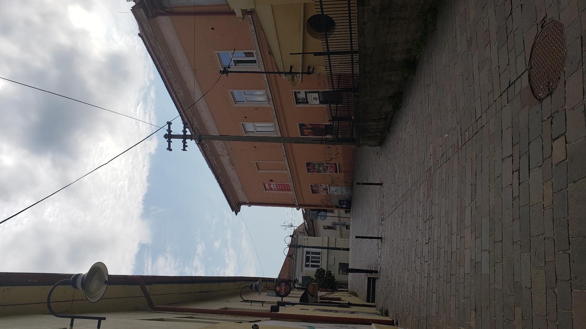The image depicts a cobblestone street in the city of Prešov, Slovakia. The street is lined with buildings, some of which appear to be old. On the left side of the street, there is a building with a yellow facade and a street lamp attached to the wall. Further down the street, there is another building with a cream-colored facade and arched windows. On the right side of the street, there is a prominent orange building with several windows and some posters on its facade. A black metal fence runs along the sidewalk in front of this building. The sky is partly cloudy, with a mix of blue sky and white clouds. There are power lines and utility poles visible above the buildings. The overall scene suggests a quiet, urban environment.