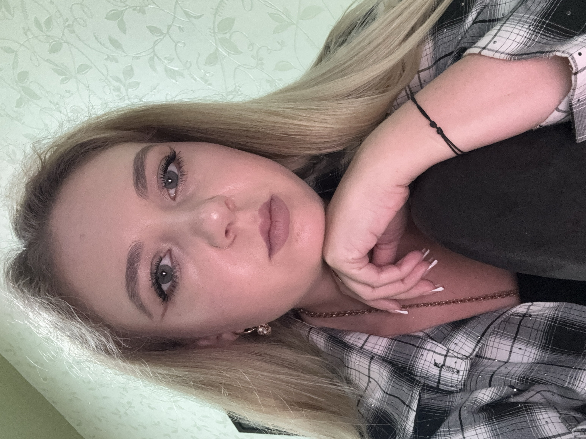 A close-up, head and shoulders shot of a young Caucasian woman with blonde hair and blue-green eyes, leaning her chin on her hand. She is wearing a black and white plaid shirt with rhinestones. She has on makeup, including eyeliner and lip gloss. The background is a light green wallpaper with a floral pattern. The image is lit with artificial light, creating a warm tone. No specific location in Makiivka, Ukraine can be determined from the image.