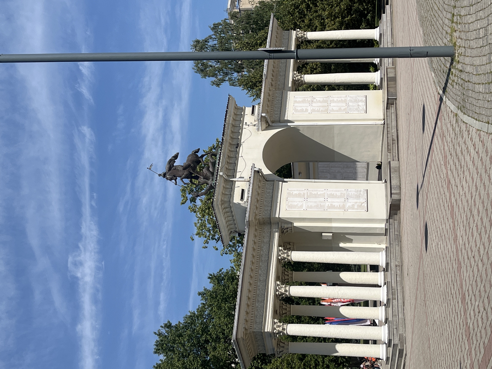 This is a medium shot, eye-level, outdoor photograph taken on a sunny day with a bright blue sky and wispy white clouds. The central subject is a white neoclassical monument with a large archway, flanked by colonnades of white pillars. Atop the monument is a dark bronze sculpture of a figure on horseback, appearing to be engaged in combat, possibly Saint George and the Dragon, though the dragon is partially obscured.
On the left side of the monument, a row of white pillars with Corinthian capitals supports a decorated entablature. Behind these pillars, several flags are visible, including red, white, and blue ones. To the right of the archway, another set of similar pillars and entablature are present. The archway itself has an inscription or list of names on its interior walls, as do the flat sections of the walls behind the colonnades. The monument stands on a raised platform, accessed by steps.
The ground in front of the monument is paved with reddish-brown and grey interlocking bricks, forming a patterned walkway. A dark grey lamppost stands in the foreground on the right, casting a shadow onto the pavement. In the background, lush green trees surround the monument, and a building with light-colored walls is visible further in the distance. A few people are present on the far left, partially obscured by the colonnade, with one person appearing to be on a bicycle or scooter.
The monument is likely the Victory Arch or Triumph Arch in Krasnodar, Russia, commemorating the liberation of the city during World War II. The sculptural element on top is commonly identified as Saint George slaying the dragon, a symbol of victory over evil, and is a prominent feature of the arch. The inscription panels likely list the names of fallen soldiers or significant events related to the city's liberation.