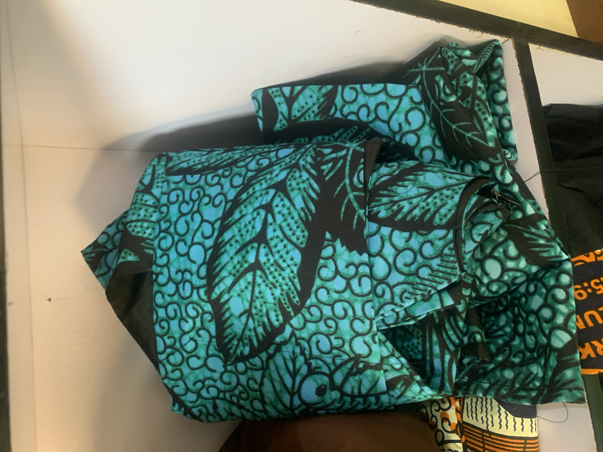 The image shows a close-up view of folded fabric with a teal and black tribal pattern, stacked on a white shelf in what appears to be a storage area. The fabric has a repeating design of leaves and swirls. Part of another fabric with an orange and yellow geometric pattern is visible to the left. The background consists of a plain white wall and shelf. There are no people or visible actions taking place. The specific location of Jauro Yinu, Nigeria, cannot be determined from the visual information provided.