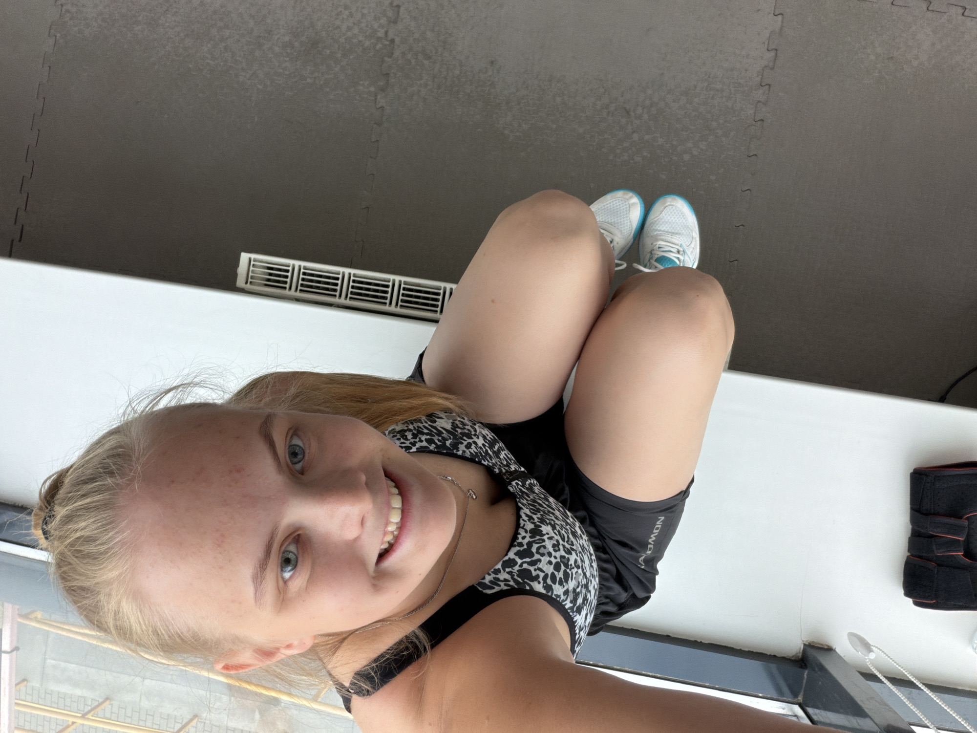 A woman is posing indoors. She is wearing a black and white sports bra, black athletic shorts, and white sneakers with blue accents. The shorts have the word "NOWON" visible on the side. She is smiling and looking directly at the camera. The background includes a grey tile floor, a white ledge, and an air vent. There is an open window or balcony door in the background. A black knee brace and a silver necklace are also visible. The location cannot be determined based on the image alone.