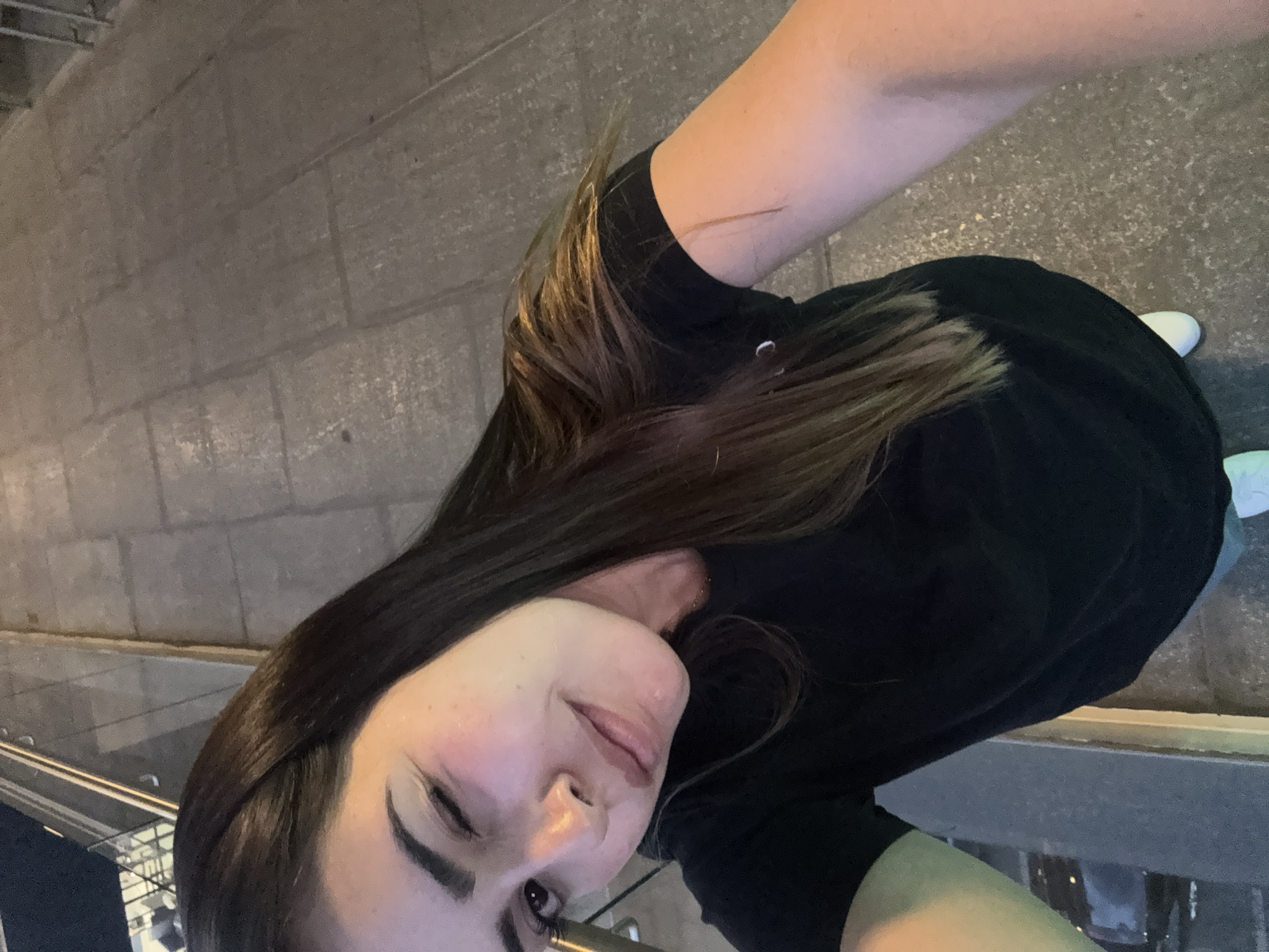 An overhead selfie captures a person from the chest up, looking down towards the camera with a subtle, winking or smiling expression. The subject has long, dark brown hair with lighter highlights, which falls over their left shoulder and arm. They are wearing a simple black t-shirt or top. Part of their left eye, nose, and mouth are visible, with rosy cheeks suggesting blush or warm lighting. White athletic shoes are partially visible in the bottom right corner of the frame.
The background shows a paved ground composed of rectangular grey blocks, indicative of a sidewalk or pedestrian area. In the lower left, a metallic railing or barrier is visible, suggesting the edge of a raised platform or stairs. The overall lighting is dim, suggesting it is nighttime or late evening, with some blurred artificial lights or reflections in the distant background. The scene is set in an urban or semi-urban environment in San Bernardino Tlaxcalancingo, Mexico. There is no visible text in the image.