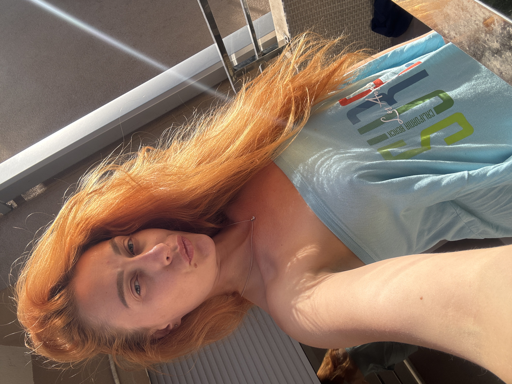 A woman with long, bright red hair is taking a selfie on a balcony in Budva, Montenegro. She is wearing a light blue off-the-shoulder t-shirt with the words "California Beach Los Angeles" printed on it in colorful block letters. The sunlight illuminates her face and hair, casting shadows on her skin and the t-shirt. The background shows a railing, a building with shuttered windows, and a glimpse of a table.