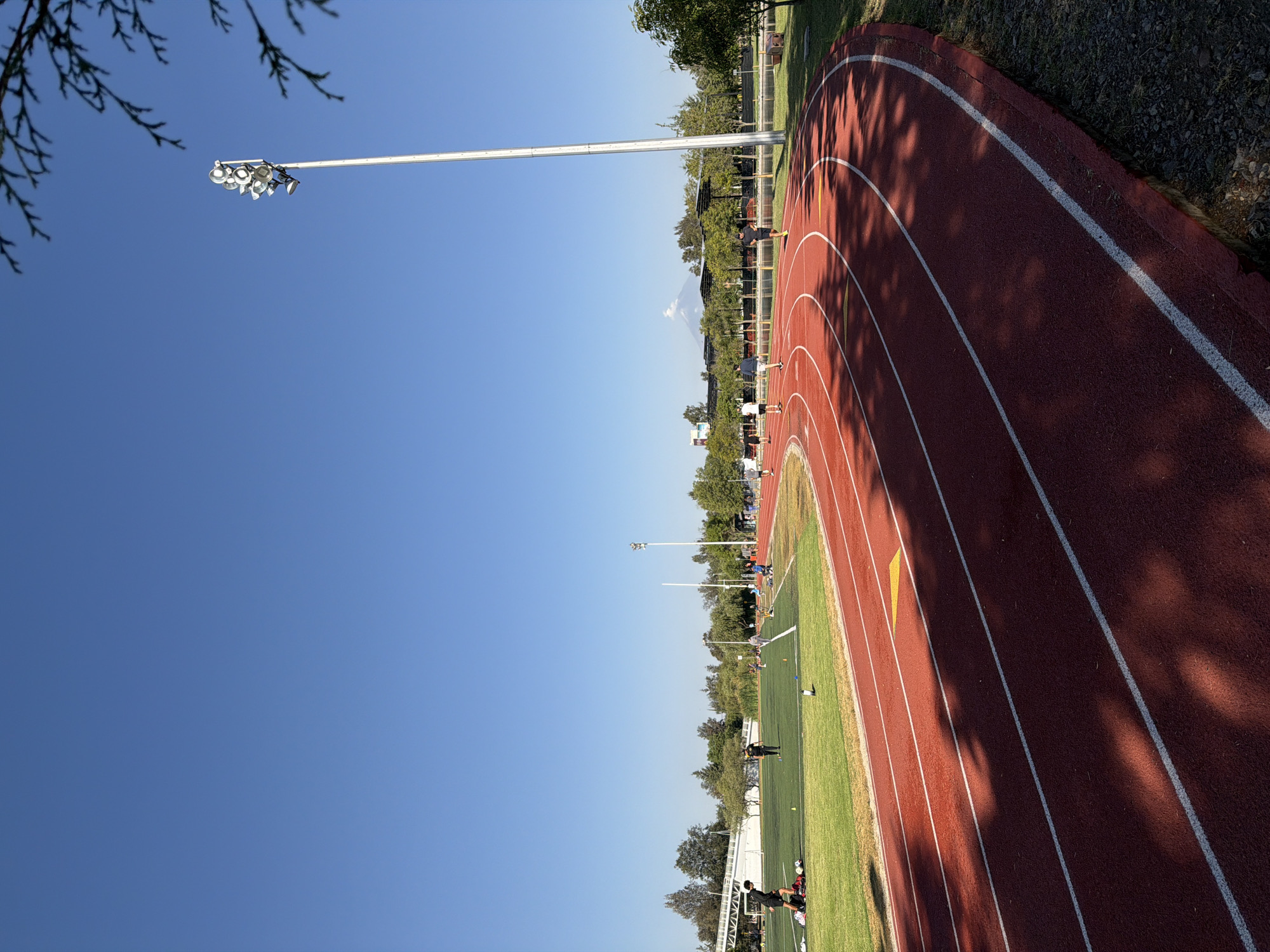 The image depicts an outdoor athletic complex under a clear, bright blue sky, likely in Puebla, Mexico, given the prominent distant mountain. The scene is oriented vertically in the original image but described here as if correctly rotated.
In the foreground, the branches of a tree frame the upper left corner, while a tall, slender white light pole with multiple fixtures extends into the sky from the left.
The midground features a vibrant red synthetic running track with distinct white lane markings and some yellow triangular markers. Strong shadows from surrounding trees and possibly the light poles are cast across the track, indicating daytime with the sun at an angle. Inside the track is a lush green grassy field, appearing to be a football or soccer pitch.
Several individuals are present on both the track and the field, engaged in what appears to be sports training or recreational activities. One person is jogging on the track, while others are standing or moving on the field, with some sports equipment like cones and balls visible.
Beyond the field, a line of green trees separates the complex from further structures or buildings. In the far distance, a majestic mountain, possibly Popocatépetl or Iztaccihuatl, rises against the horizon, its peak partially obscured by clouds or snow. The weather is clear and sunny. No readable text is visible within the image.
