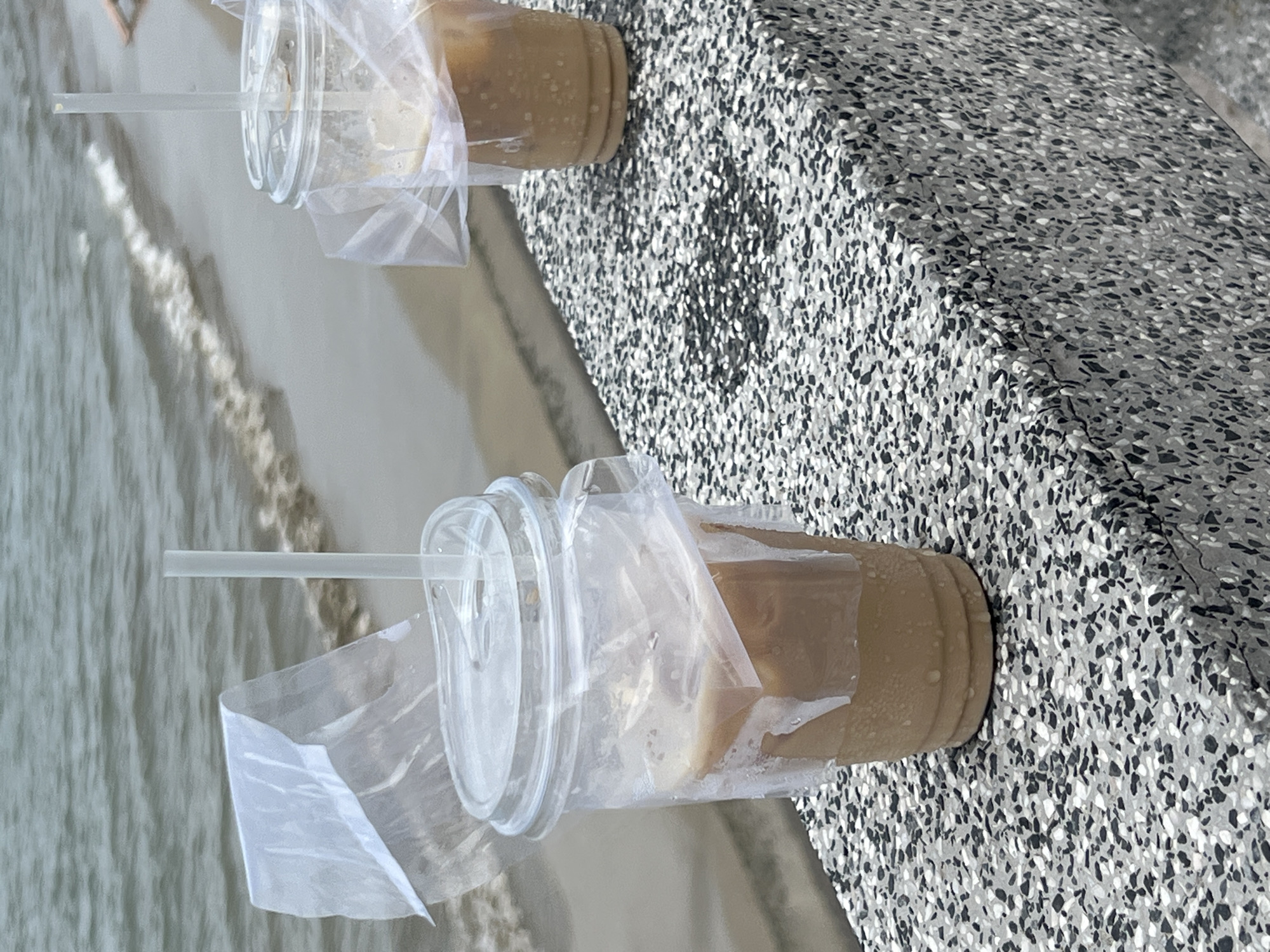 The image displays two clear plastic cups, each with a lid and straw, containing a light brown, milky beverage, likely iced coffee. White plastic bags are wrapped around the cups. These drinks are placed on a speckled gray concrete ledge or wall, which occupies the right side of the frame. In the background, the ocean stretches out with gentle waves and some whitewash. A person's leg or foot is partially visible in the top left, suggesting someone is walking along the sandy edge of the water. The scene depicts a leisurely moment by the sea, perhaps enjoying refreshments on a beach in Bac Nha Trang Ward, Vietnam. The lighting indicates it is daytime. No text is visible in the image.
