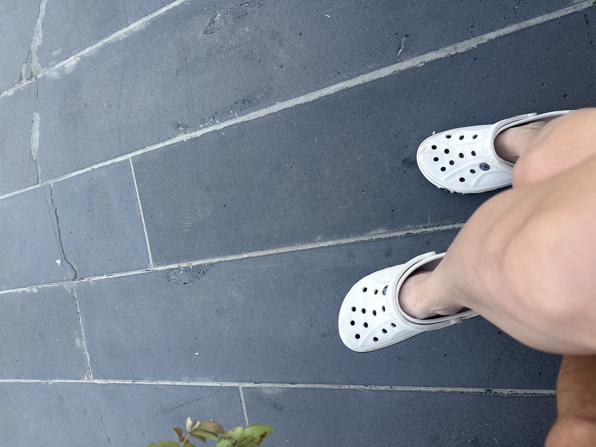 A pair of white Crocs are visible on a person's feet, standing on a dark tiled surface. One Croc is on the left foot, and the other is on the right. The tiles are rectangular and laid in a pattern, with lighter grout lines separating them. A plant with green and brown leaves is partially visible in the upper left corner of the image. The scene appears to be outdoors, possibly in a public space. The image does not provide any specific details about the city of Gyumri, Armenia, or any identifiable landmarks.