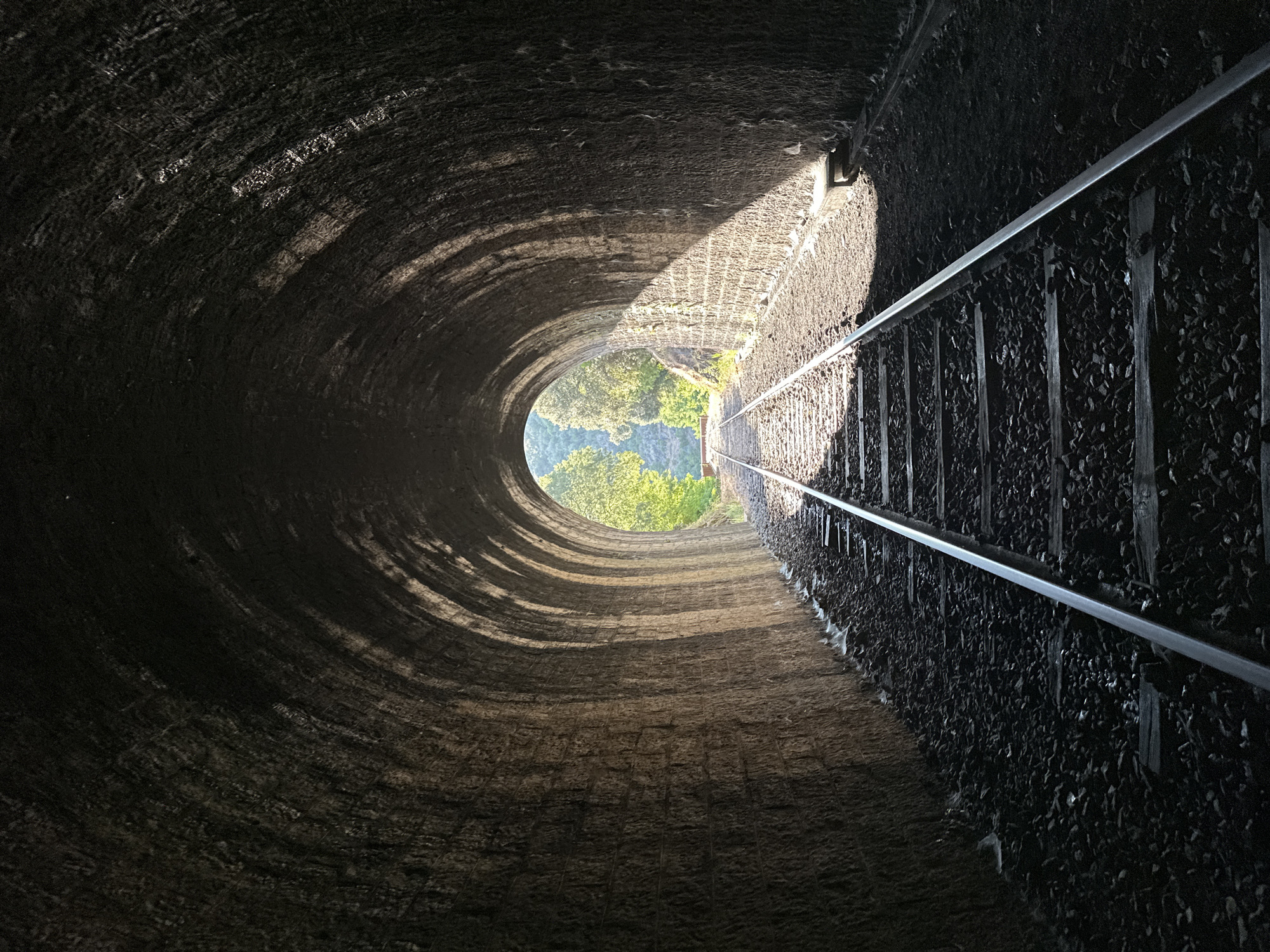 This is a photograph taken inside a stone railway tunnel in Générargues, France. The tunnel has a high, arched ceiling constructed of large, irregular stones. The walls are rough and textured, showing evidence of age and weathering. Two sets of railway tracks run down the center of the tunnel, disappearing into the darkness. The tracks are made of metal rails laid on wooden sleepers, with ballast of dark gravel.
Sunlight streams into the tunnel from the opening at the far end, creating a dramatic contrast between the illuminated exterior and the shadowed interior. The light casts strong shadows on the tunnel walls, highlighting the arched construction. Outside the tunnel, lush green trees and foliage are visible, suggesting a natural, possibly rural, environment. The overall impression is one of a historical and atmospheric location.