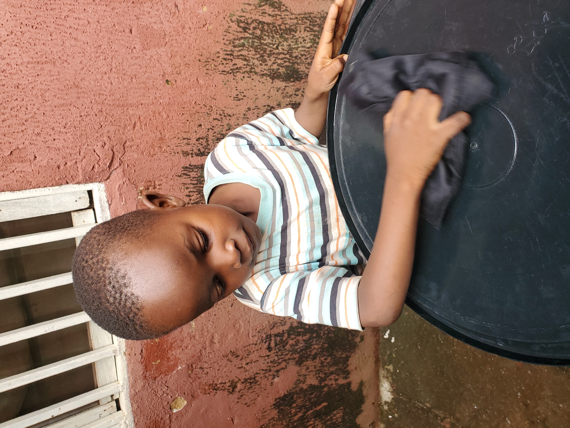 A young Black child wearing a horizontally striped t-shirt is wiping the surface of a large, black circular object with a dark cloth. The child is looking down and appears focused on the task. Behind the child, a textured reddish-brown wall is visible, with some streaks of dark discoloration. Above the wall, a white-barred window or grate is present. The ground appears to be a wet, concrete surface. The scene is outdoors, possibly in a residential area in Jos, Nigeria.