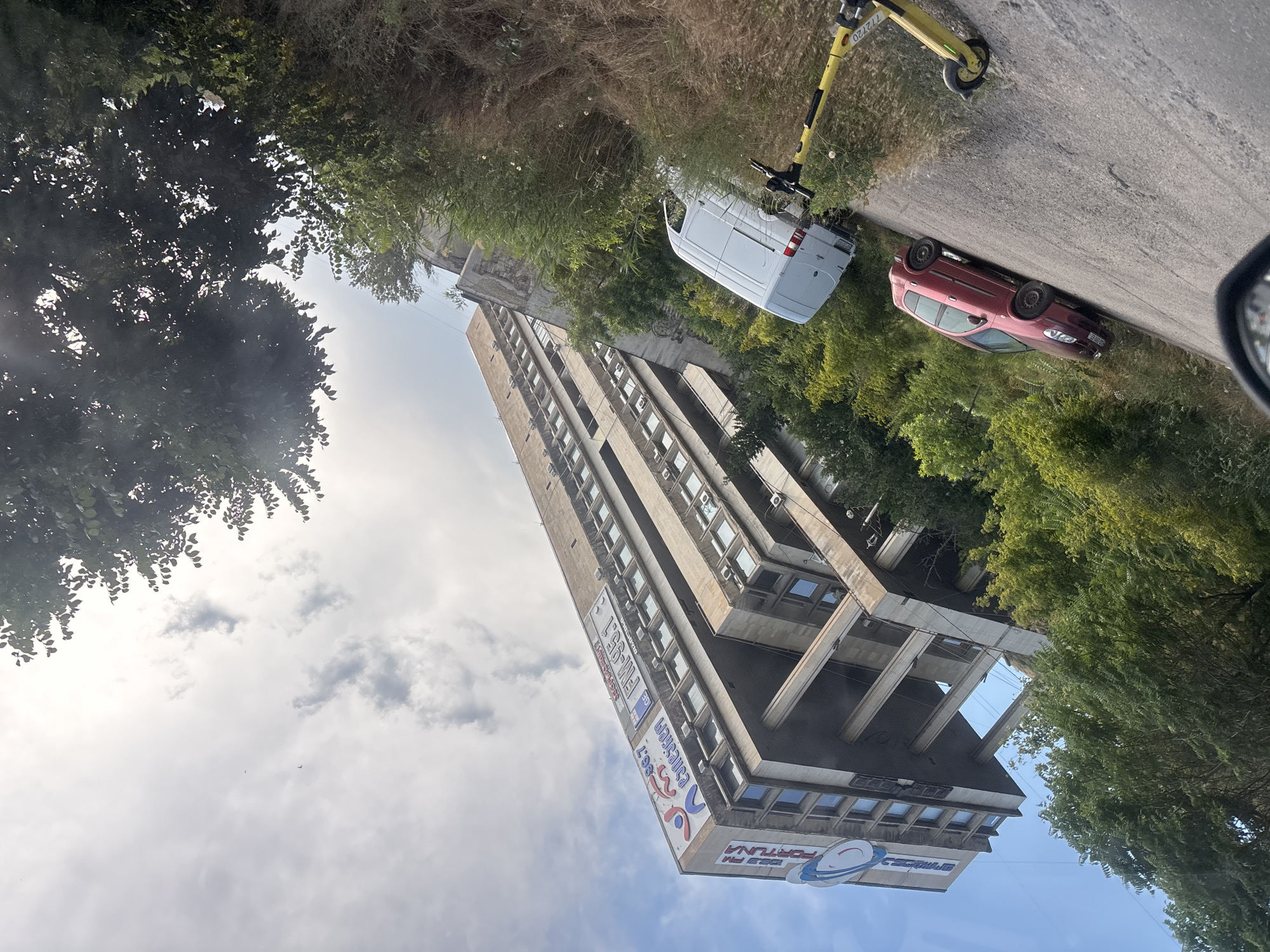 The image depicts a street scene in Tbilisi, Georgia, with a tall, Brutalist-style apartment building dominating the foreground. The building, made of concrete, features a repetitive pattern of windows and external structures. Several large advertisements are visible on the side of the building, including one with "FM-95.1" and another with "FORTUNA".
To the right of the building, a red car is parked on the side of a paved road, and further up the road, a white van is also parked. A yellow electric scooter is propped up on the roadside, near some dry grass and greenery. Lush green trees surround the area, with some foliage encroaching into the frame from the left and right. The sky is partly cloudy. The overall impression is of an urban environment with elements of nature, possibly during daylight hours, with no clear indication of a specific event or activity taking place. The composition suggests the photograph was taken from a moving vehicle.