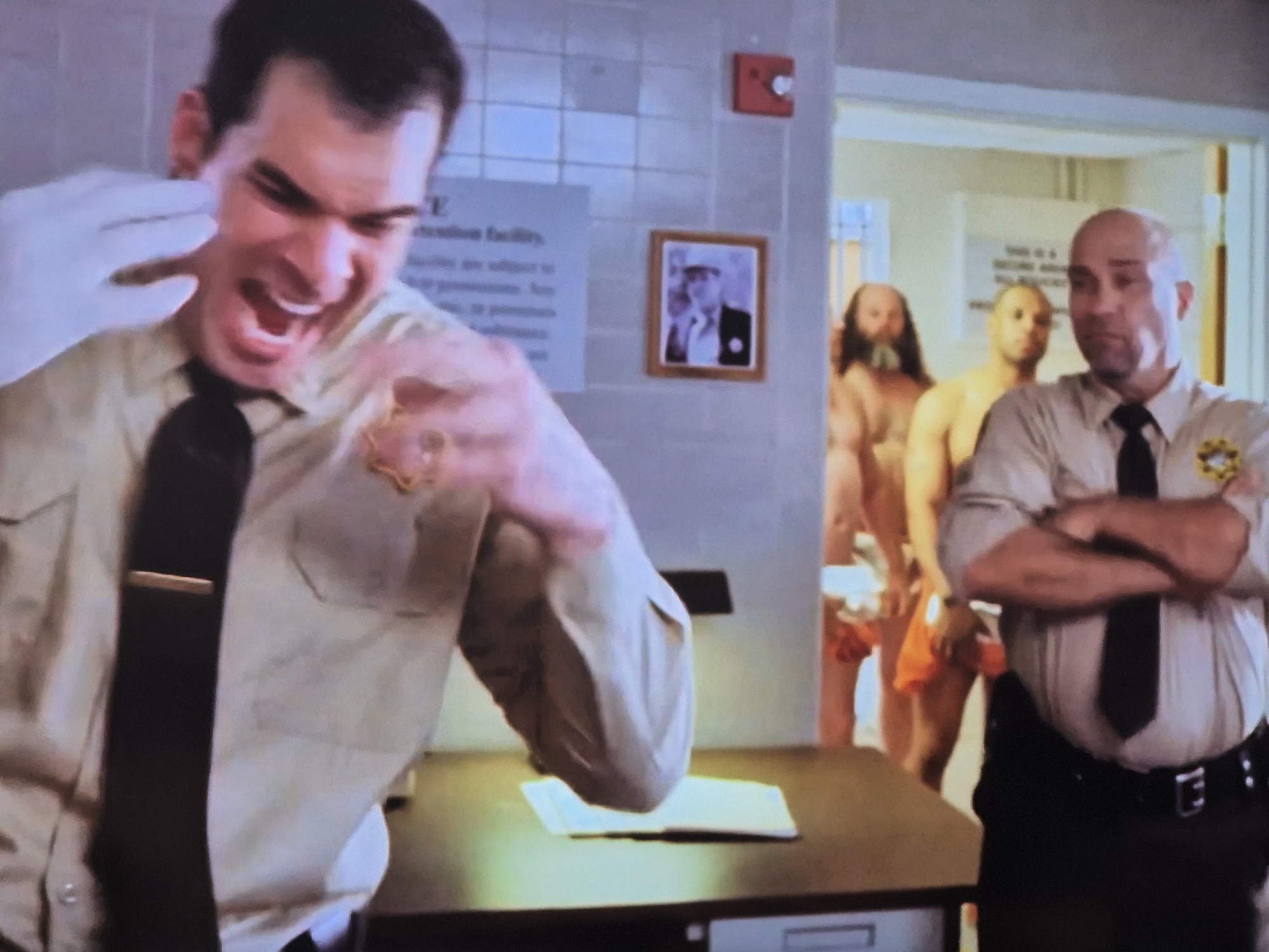 A man in a light brown uniform shirt and black tie with a badge on the left breast pocket is in the foreground, his mouth wide open in a scream and his right hand reaching out with a white glove. Behind him, to the right, is a bald man in a similar uniform and crossed arms. In the background, through a doorway, several naked men stand in a hallway. On the wall behind the man in the foreground is a framed picture of a man in a hat and suit, and a red fire alarm. The environment appears to be an indoor facility, possibly a detention center or prison.
