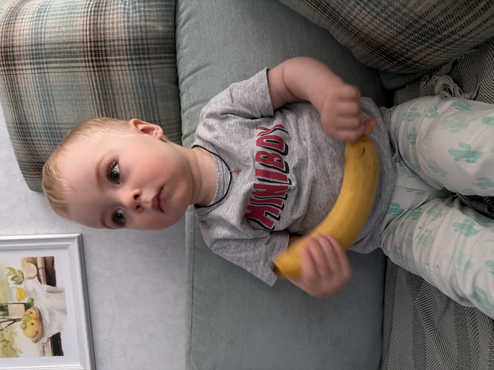 A young child lies on a light green sofa, holding a banana. The child is fair-skinned with blond hair, wearing a gray t-shirt with a red and gray design and white pants patterned with light green cacti. The child is also wearing a dark necklace with a small cross. The sofa has a patterned pillow behind the child's head and a patterned blanket to the child's right. A framed picture hangs on a light gray wall to the left of the sofa. No information is available to connect this image to the city of Votkinsk, Russia.