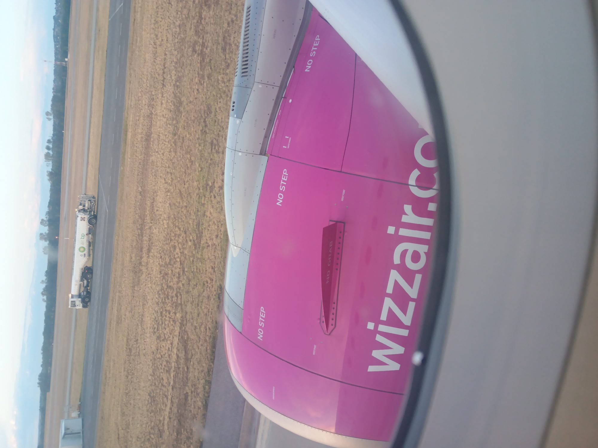 The image shows a close-up view of a Wizz Air airplane engine from inside the cabin, looking out through a circular window. The engine cowling is painted bright pink and features the airline's logo and "NO STEP" markings. A red safety cover labeled "NO GRAB" is visible on the engine. In the background, an airport tarmac is visible with a fuel truck with "air bp" branding. The sky above is overcast with a pale blue hue. The scene appears to be at an airport, likely Budapest Ferenc Liszt International Airport, given the Wizz Air branding.