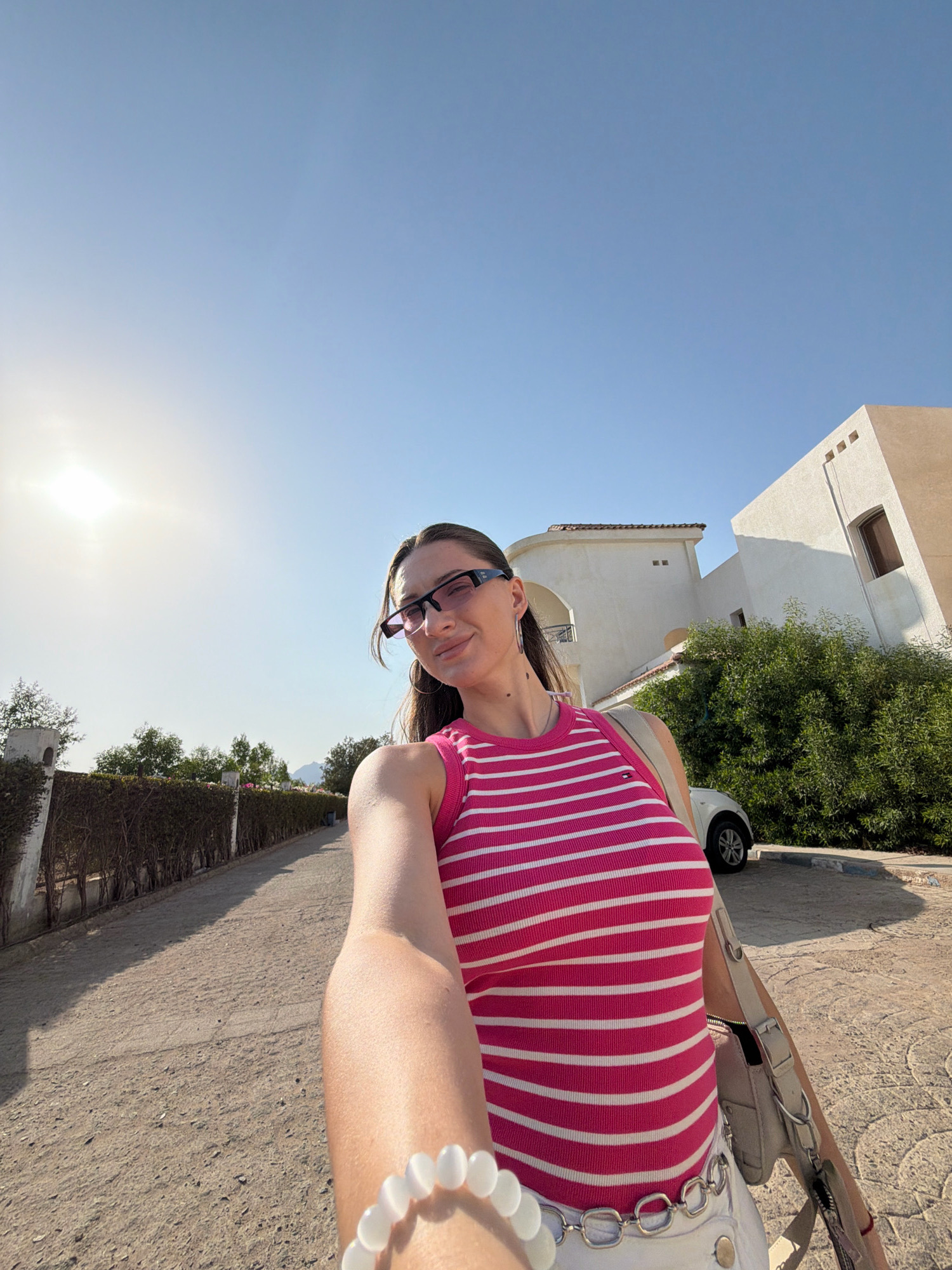 A woman is taking a selfie in Sharm El Sheikh, Egypt. She is wearing a pink and white striped tank top, white shorts, and glasses. She is also wearing a bracelet and a purse. She is standing in front of a building and some trees. The sun is shining brightly in the sky.