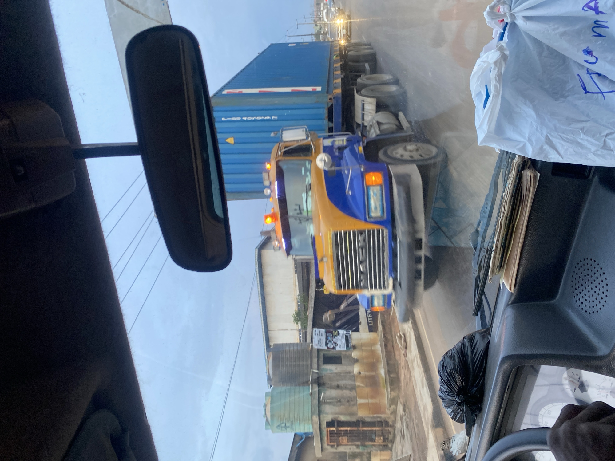 A two-axle blue and yellow Mack semi-truck, hauling a blue shipping container, drives on a wet, paved road. The truck's hazard lights are illuminated. The scene is viewed from inside a vehicle, with the dashboard, steering wheel, and a black trash bag visible in the foreground. The sky is overcast, and power lines are visible in the background. In the distance, other vehicles are on the road. Buildings, including structures with water tanks, are visible on the left side of the road. The image was taken in the city of Ikorodu, Nigeria.