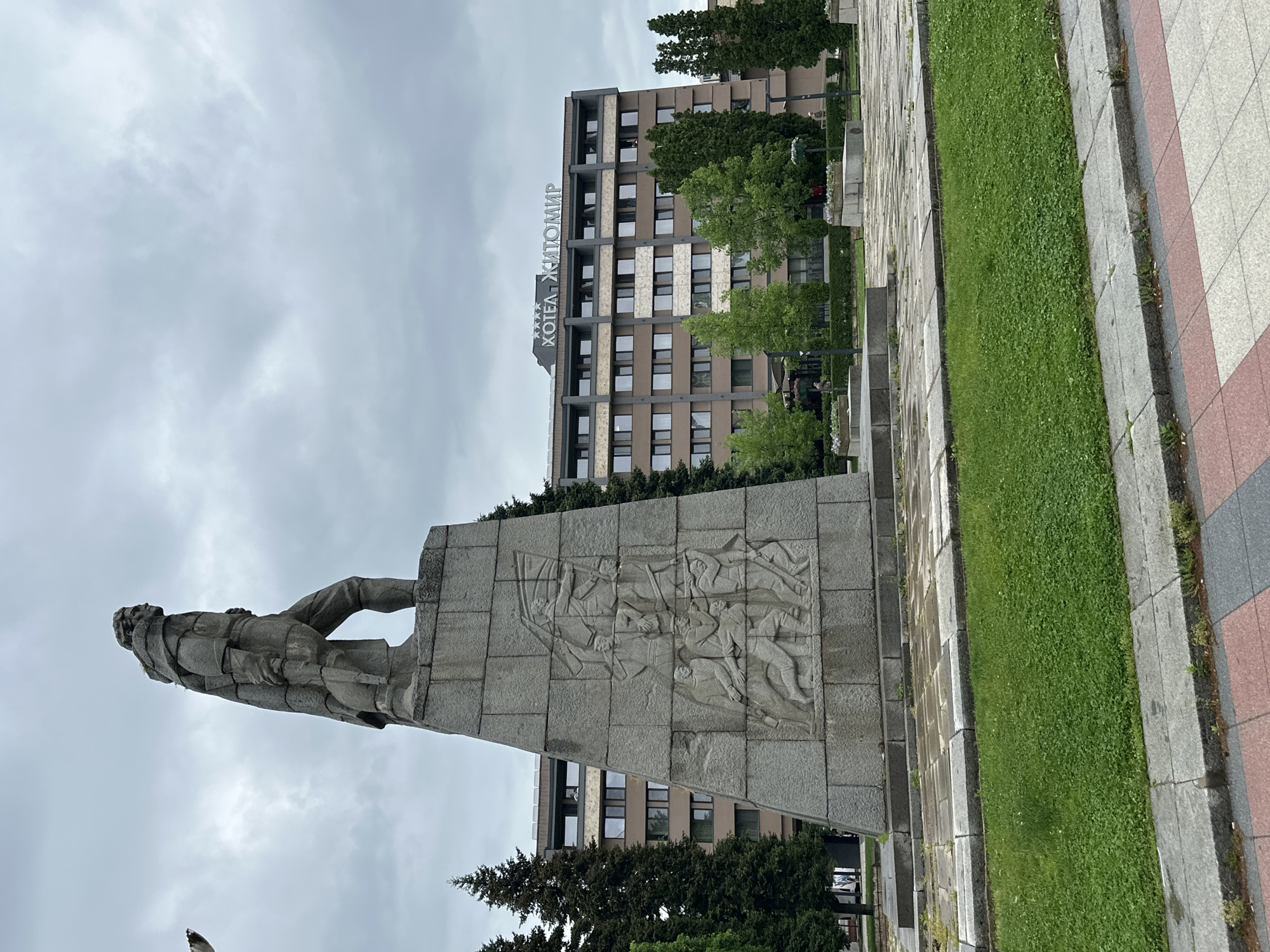 The image shows a concrete monument in Montana, Bulgaria, with a statue of a man at the top. The side of the monument features a relief carving depicting several figures. Behind the monument is a multi-story building with the Cyrillic text "ХОТЕЛА ЖИТОМИР" on its facade. The sky is overcast, and the area around the monument includes green grass, paved walkways, and trees.