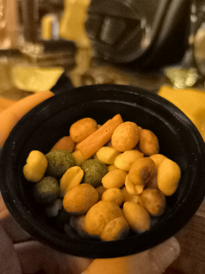 A close-up shot captures a black bowl filled with an assortment of colorful snacks. The primary contents include numerous orange-coated nuts or peanuts, alongside some green-coated nuts or peas, and lighter yellow/beige nuts, likely roasted peanuts. Additionally, several elongated orange stick-shaped snacks, resembling mini carrot sticks or cheese straws, are visible. The snacks are in sharp focus, showcasing their varied shapes and vibrant hues of orange, yellow, and green.
A light-skinned hand is partially visible at the bottom left, holding the bowl, suggesting the snacks are being prepared for consumption or are currently being eaten. An additional bright orange object, possibly another food item, rests beside the bowl on the left. The background is softly blurred with warm tones of yellow, orange, and brown, indicating an indoor environment, though specific details are indistinct. The overall scene suggests a casual snacking occasion. No text is visible in the image. This event is taking place in Palaio Faliro, Greece.