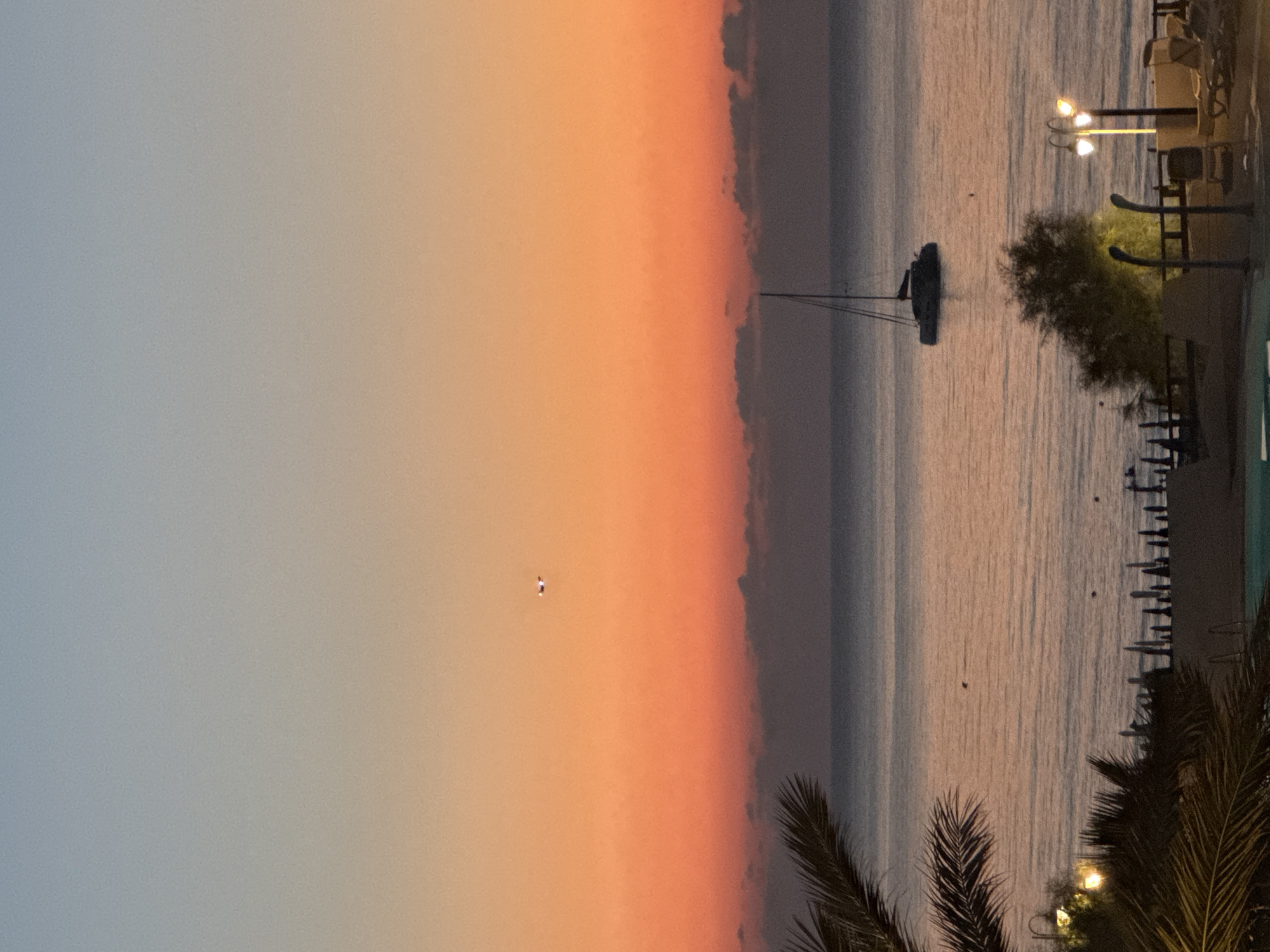 At dusk, a catamaran sails on calm waters off the coast of Isola delle Femmine, Italy. The sky transitions from a pale blue to a vibrant orange and pink gradient. A distant airplane is visible against the colorful sky. Palm fronds frame the left side of the scene, while beach umbrellas, lounge chairs, and lampposts are visible along the shoreline in the foreground. The water reflects the warm hues of the sunset.