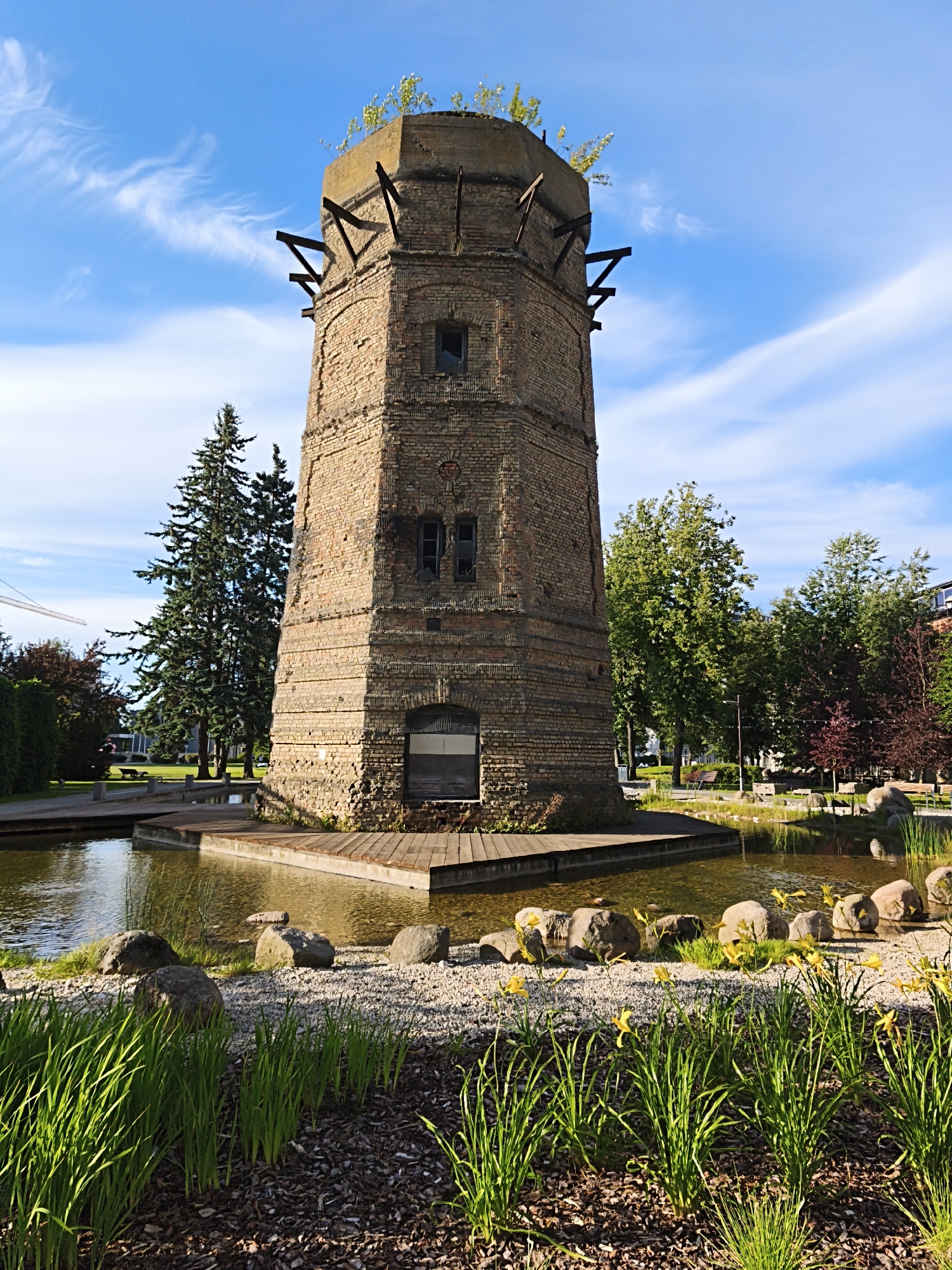 The image shows a round, brick tower with a concrete top and wooden supports protruding from the upper part. The tower is surrounded by a moat with wooden decks and is situated in a park-like setting with trees, grass, rocks, and yellow flowers. The sky is blue with wispy white clouds. In the background, there are more trees, buildings, and a crane. This is the historic Kalamaja water tower in Tallinn, Estonia, a former industrial building that has been repurposed as a public space.