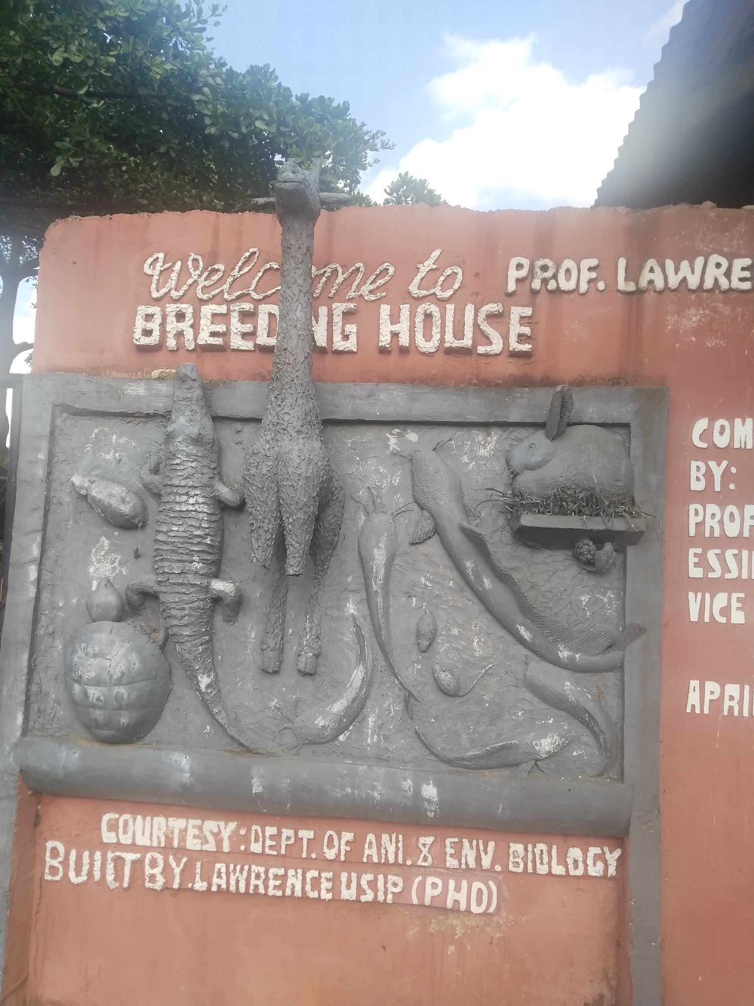 The image depicts a concrete sign for the "Prof. Lawrence Breeding House," likely located in Uyo, Nigeria. The sign features a raised relief sculpture of various animals, including a turtle, crocodile, giraffe, fish, and rabbit. The sign also includes text indicating that the sign was "Courtesy: Dept. of Ani. & Env. Biology" and "Built by Lawrence Usip (PhD)." The background includes trees and a building. The overall condition of the sign appears aged.