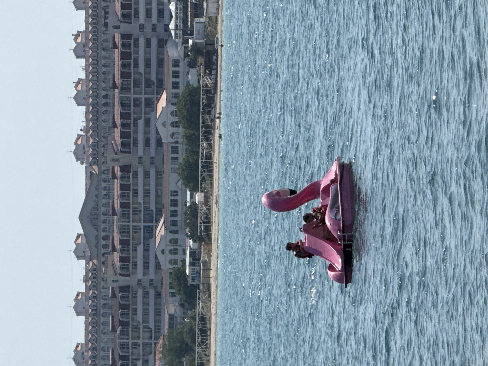 A pink flamingo-shaped paddleboat with two young passengers is on the choppy, blue water of the Black Sea. The boat is positioned in the foreground, with its beak and head pointing to the right. The passengers, wearing life vests, are seated in the center of the boat. In the background, a long, multi-story building complex with red-tiled roofs lines a sandy beach. Several trees are visible between the beach and the building. The sky is clear and pale. The scene appears to be a sunny day at a seaside resort. The location is likely a beach in Sevastopol, Ukraine, given the architecture of the buildings which are typical of resort towns in the region.