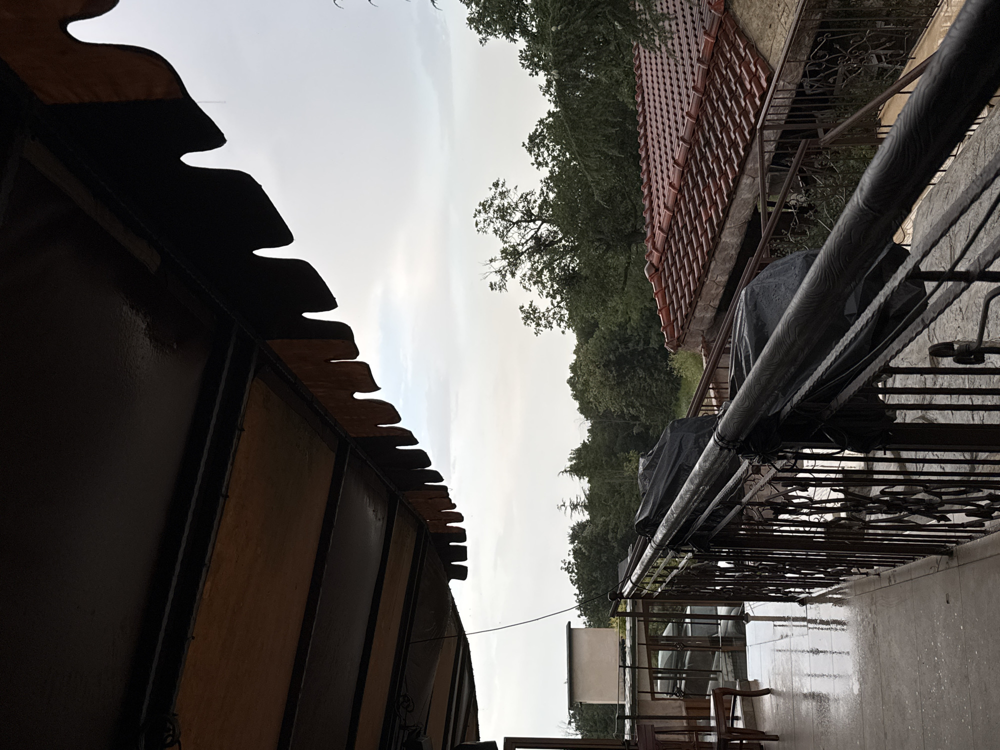 The image depicts a balcony with a wet tiled floor, indicating recent rain. A dark metal railing runs along the balcony, covered in places by black tarps. Beyond the railing, a red-tiled roof of a building is visible, surrounded by lush green trees. The sky is overcast and grey. A small, light-colored structure is visible in the background amidst the trees. The perspective suggests a view from a slightly elevated position, possibly from an upper floor or hillside. The scene appears to be an outdoor residential area.