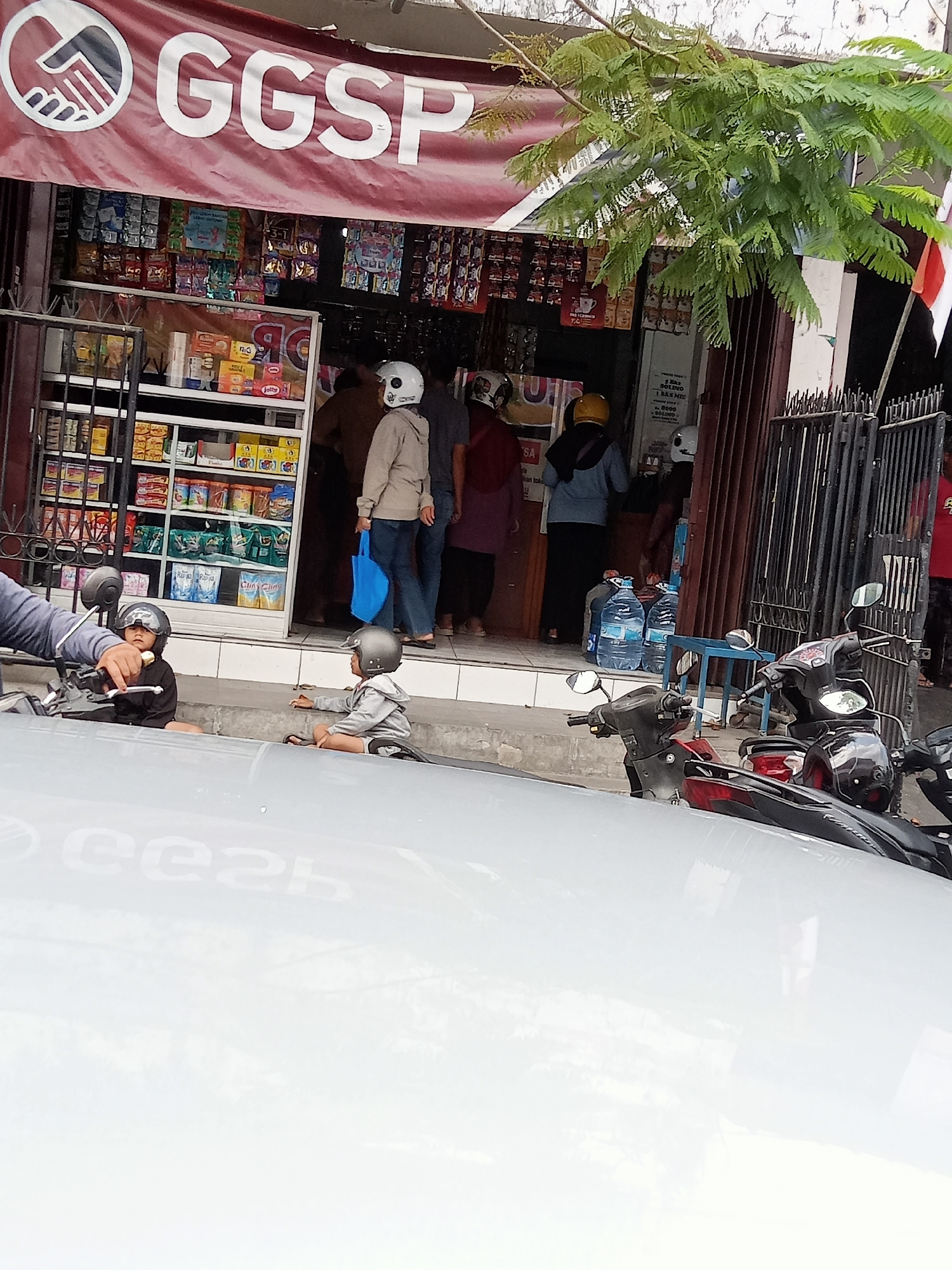 This image captures a street scene in Tulungagung, Indonesia, focusing on a small storefront with a large banner displaying "GGSP." The storefront appears to be a convenience store or small grocery, with shelves stocked with various packaged goods visible through the glass doors.
In the foreground, two children, both wearing helmets, are seated on a raised concrete area, possibly a sidewalk or a step. One child is on the left, wearing dark clothing and looking towards the viewer. The other child, on the right, is wearing light gray clothing and appears to be reaching out towards something on the ground. A motorcycle is partially visible on the left, with a hand on the handlebars, suggesting it is parked or being held.
Behind the children, several adults are visible entering or standing near the storefront. One person in a light brown jacket and helmet is walking away from the store, carrying a blue bag. Another person in a darker jacket and a yellow helmet is visible near the entrance. The storefront itself has metal grates and glass panels, typical of shops in the area.
To the right of the storefront, large bottles of water are stacked on a small blue table. Further to the right, a dark metal fence or gate is present, with a glimpse of a red and white flag at the top. The overall environment appears to be an outdoor, daytime setting with some overhead foliage from a tree casting shadows. The ground in the immediate foreground is a reflective white surface, possibly a car hood, obscuring the lower part of the scene.