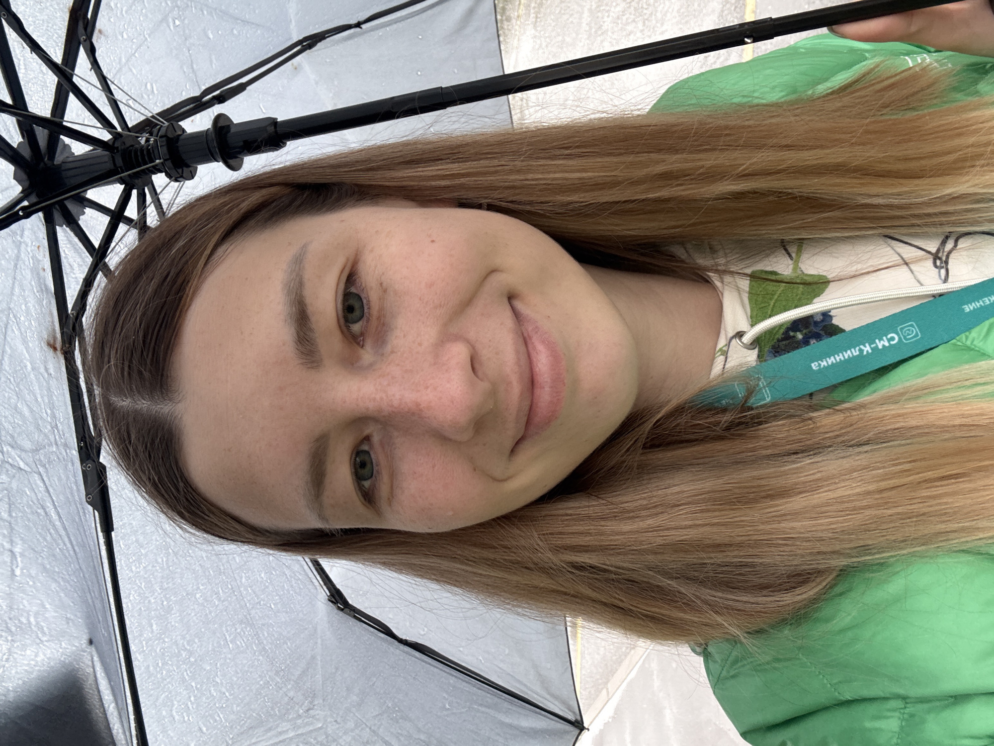 A woman is holding a black umbrella on a rainy day. She is wearing a green jacket and a lanyard with Russian text visible: "СМ-Клиника." This indicates a location possibly associated with the SM-Clinic healthcare network, which has branches in Moscow, Russia. The scene is likely outdoors, given the umbrella and rainy weather. The woman is smiling at the camera.