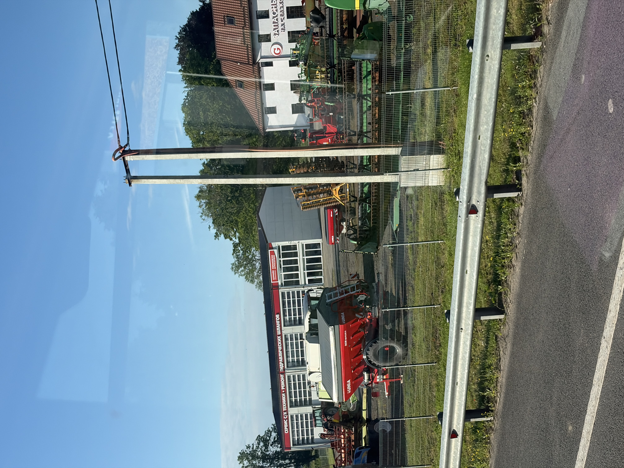 The image depicts an outdoor scene, likely a commercial property, with agricultural machinery and buildings. In the foreground, a metal guardrail lines the edge of an asphalt road. Behind the guardrail, a chain-link fence encloses a grassy area with various pieces of agricultural equipment, including a red tractor, a large red seeder with the brand "UNIA" visible, and other harvesting or tilling machinery. In the background, there are two buildings. One is a long, white building with multiple garage doors, labeled in Russian with text that appears to be "СЕРВИС С/Х ТЕХНИКИ • РЕМОНТ ГИДРАВЛИЧЕСКИХ ШЛАНГОВ" (Service of agricultural machinery, repair of hydraulic hoses). Next to it is a white building with a red tile roof and several windows, which has a red circular logo with a white "G" and Russian text that appears to say "ЗАПАСНЫЕ ЧАСТИ НА СЕЛЬХОЗТЕХНИКУ" (Spare parts for agricultural machinery). The sky is blue with some wispy clouds, and trees are visible behind the buildings. There are no people visible in the image. The overall impression is a dealership or service center for agricultural equipment.