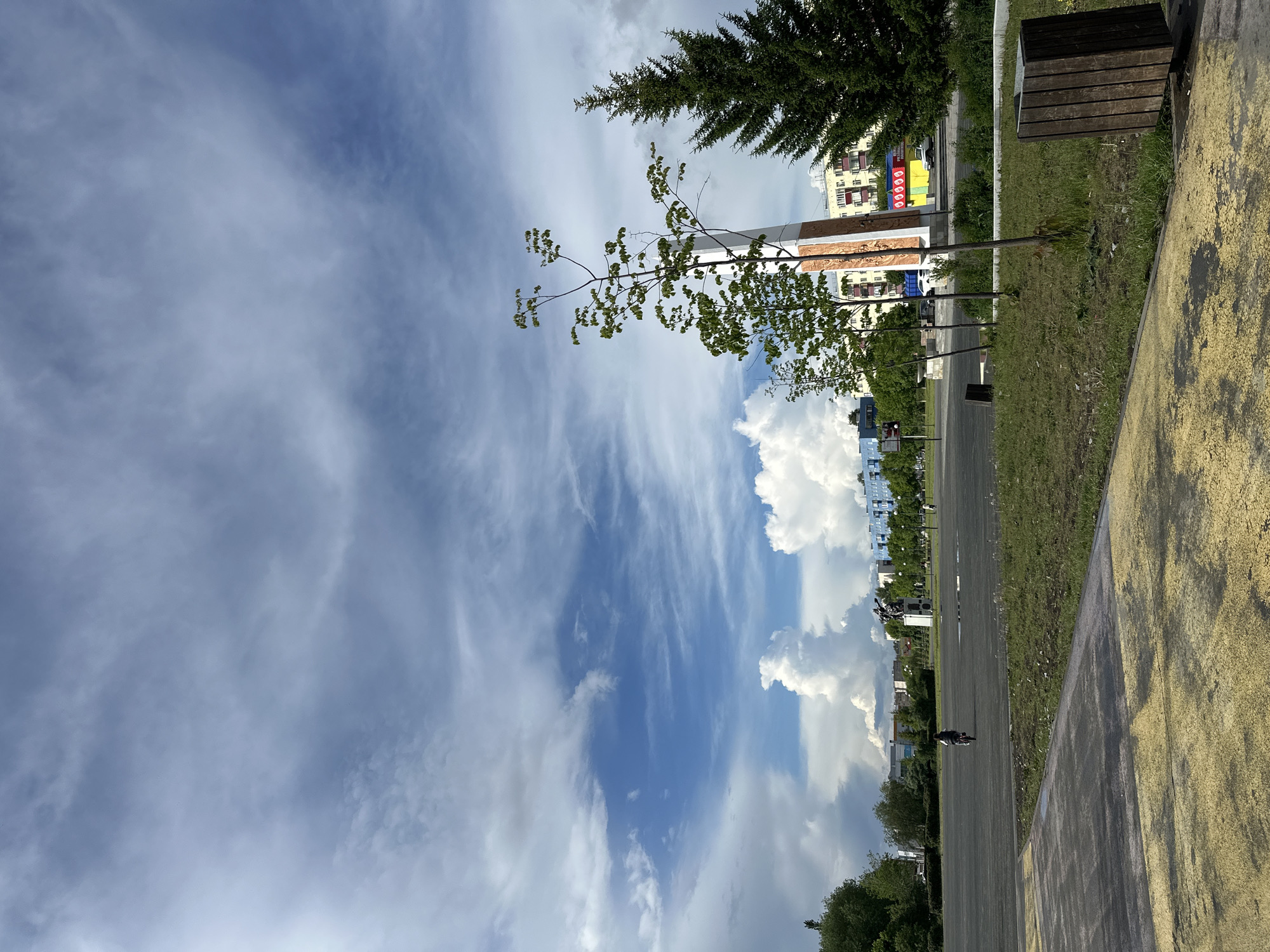 The image depicts an outdoor scene under a dynamic, partly cloudy sky, likely captured in an urban park or along a public pathway. The perspective looks down a wide, dark-paved road or path that stretches into the distance, with a grassy area and a textured yellowish-brown path running parallel to its right.
The sky dominates the upper half of the frame, showcasing a mix of wispy, high-altitude clouds resembling cirrus streaks across a blue sky, interspersed with large, fluffy white cumulus clouds, particularly concentrated on the right side. The overall lighting suggests it is daytime.
In the midground, a tall, light-colored monument or pillar with decorative carvings at its base stands prominently. On its upper section, a rectangular sign is visible, featuring five red square panels, each containing a white circle. Various trees are present, including a dark green evergreen tree near the top right and several deciduous trees with vibrant green leaves. A dark wooden bench with slatted planks is situated on the grass strip to the right.
Further down the paved path, a person is visible riding a bicycle, indicating casual recreational activity. In the background, beyond the trees and structures, several multi-story residential buildings can be seen.
The scene conveys a peaceful and common urban or suburban environment, with no explicit location cues beyond a typical park or street setting. The weather is bright but with significant cloud cover, characteristic of a pleasant day.
