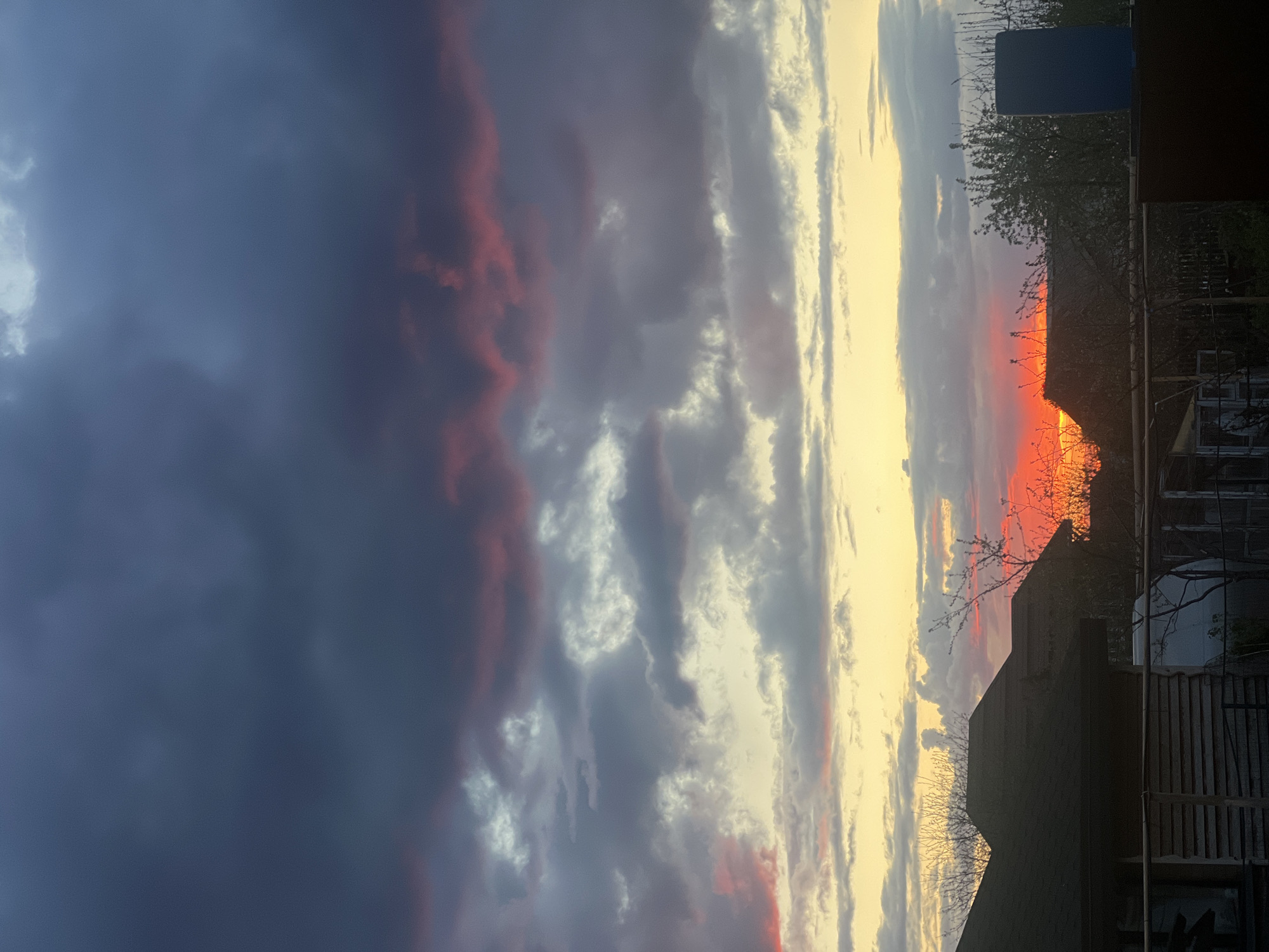 The image shows a sunset over what appears to be a residential area. A portion of a building, possibly a house or apartment block, is silhouetted against the bright, orange and yellow sky near the horizon. Trees and branches are also visible in silhouette. The sky transitions from darker, gray-blue storm clouds on the left to the bright, colorful sunset on the right, with some pink and red tinges visible in the clouds. The photo has a slight upward angle. There are no people visible. The image is possibly from Votkinsk, Russia.