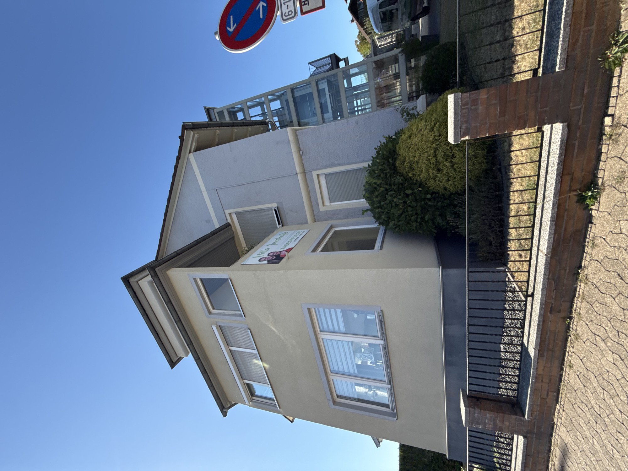 In Herzberg, Germany, a multi-story building with a light-colored facade and a grey roof stands under a clear blue sky. The building features several windows, some with roller shutters. A modern glass elevator structure is attached to the side of the building. A sign on the building reads "Villa Susanne" and "Tagespflege." Adjacent to the building, a traffic sign with red and blue circles and white arrows indicates a no-parking zone, with smaller signs below showing time restrictions and a red "H." A black metal fence with brick pillars borders the property. A green bush is visible in front of the building. The ground in front of the fence is paved with interlocking stones.