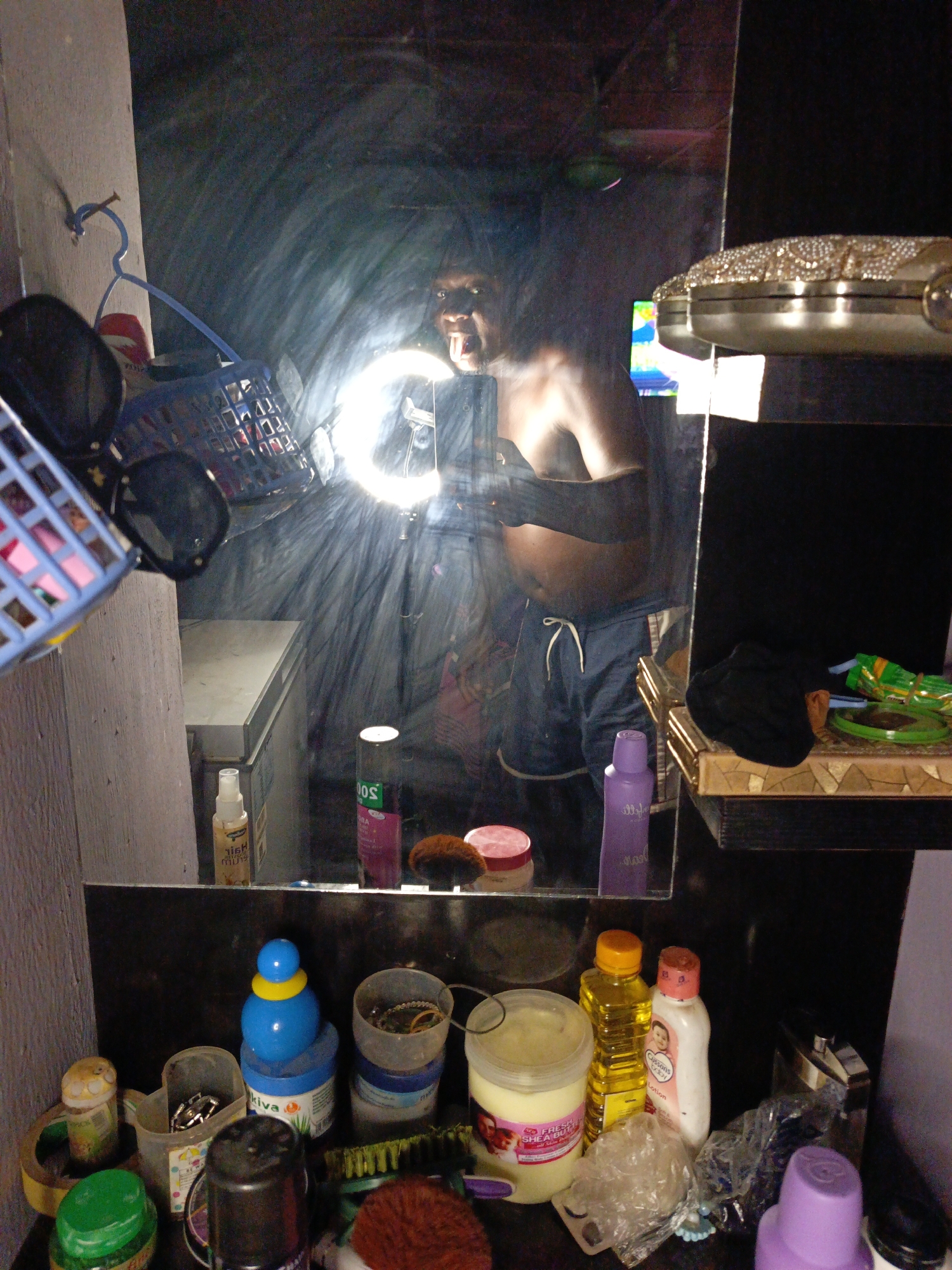 A cluttered scene in an indeterminate indoor location. A mirror reflects a person holding a cell phone with a ring light. The person is shirtless, wearing shorts and sticking out their tongue. The area in front of the mirror is filled with a variety of items, including containers of various sizes and colors, cosmetics, accessories, and a purse. A blue basket with sunglasses hangs to the left. A shelf holding black fabric and green items is located to the right of the mirror.