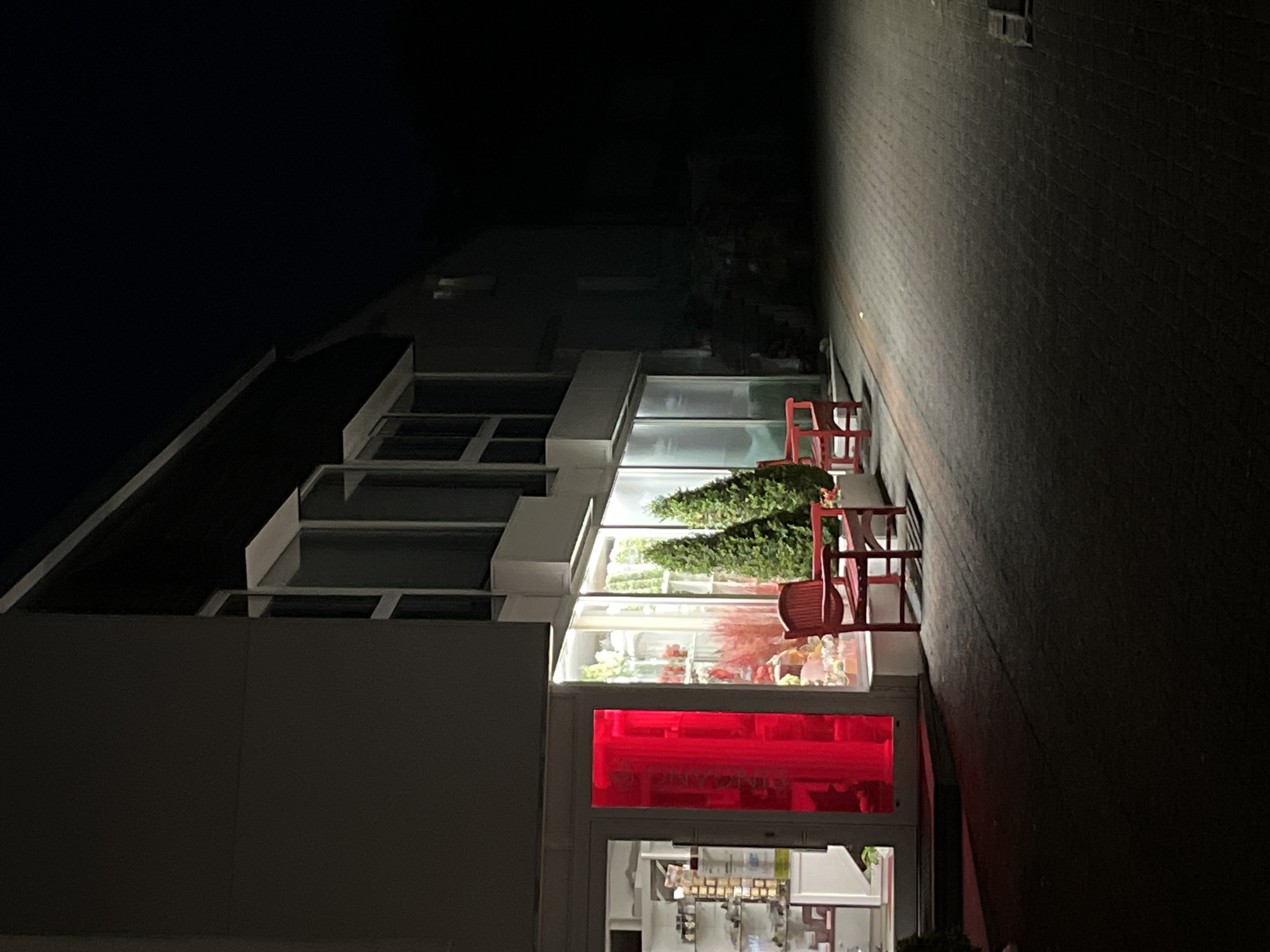The image depicts the exterior of a shop at night in Saint-Vith, Belgium. A dark, paved walkway leads up to the storefront, which features large glass windows. One window has a bright red illuminated door or panel with German text visible. Inside the shop, shelves are stocked with various items. Outside, two red wooden benches and two potted evergreen trees are visible. The building appears to be a commercial establishment, possibly a florist or gift shop, given the display inside. The lighting suggests it is dark outside, with artificial lights illuminating the shop's interior and entrance. The surrounding area is largely dark and unpopulated.