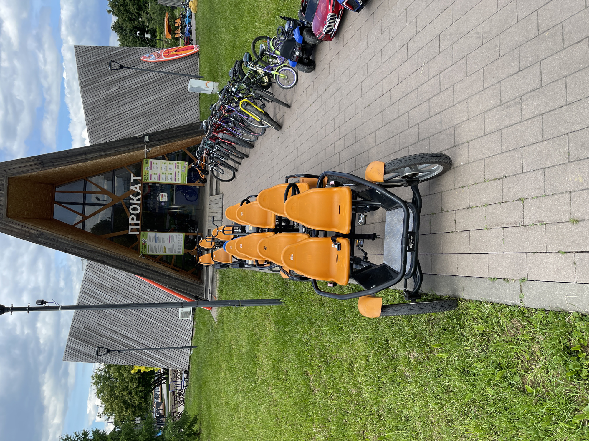 The image shows a rental shop for recreational vehicles in a park setting. The building has a modern, A-frame design with the word "ПРОКАТ" (Rental) displayed above the entrance. A variety of bicycles, including a four-person pedal-powered vehicle, are parked near the building. The environment includes a paved walkway, grass, and trees. Based on the vehicles and architectural style, the location is likely a recreational area within the city of Moscow, Russia.