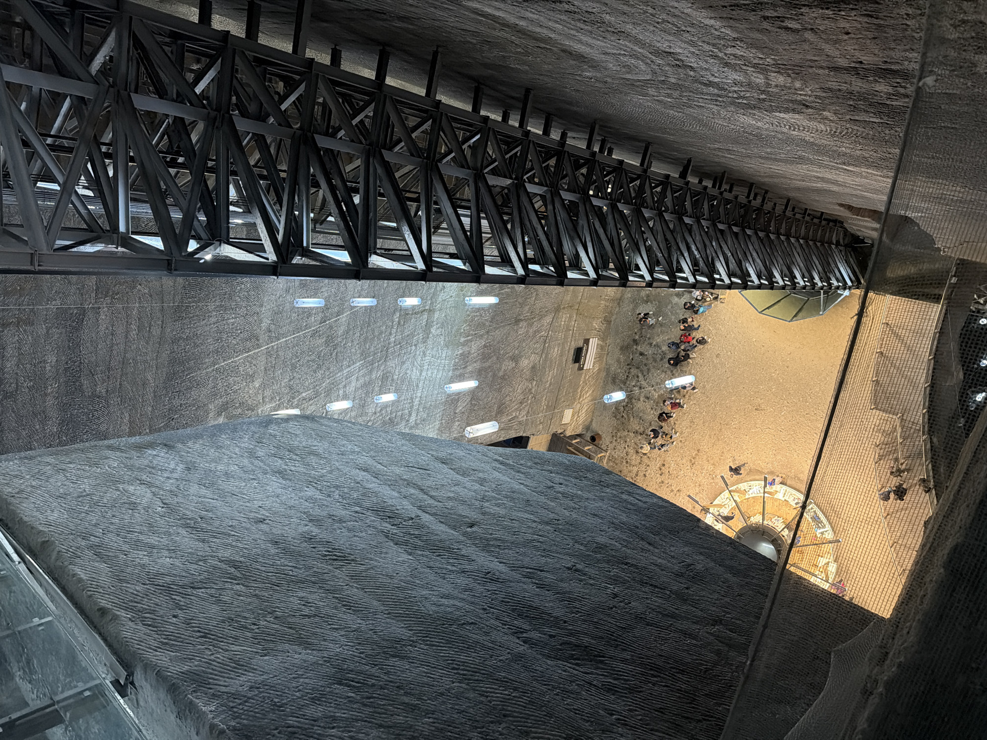A high-angle view inside Salina Turda, a salt mine in Boj-Cătun, Romania. The photo shows the rough-textured, salt-carved walls of the mine, illuminated by artificial lights. A large metal structure with a geometric pattern is attached to the upper part of the mine. Below this structure, a lower level of the mine is visible, with groups of people and a central feature, possibly a well or shaft, enclosed by a structure. A safety net is visible on the right side.