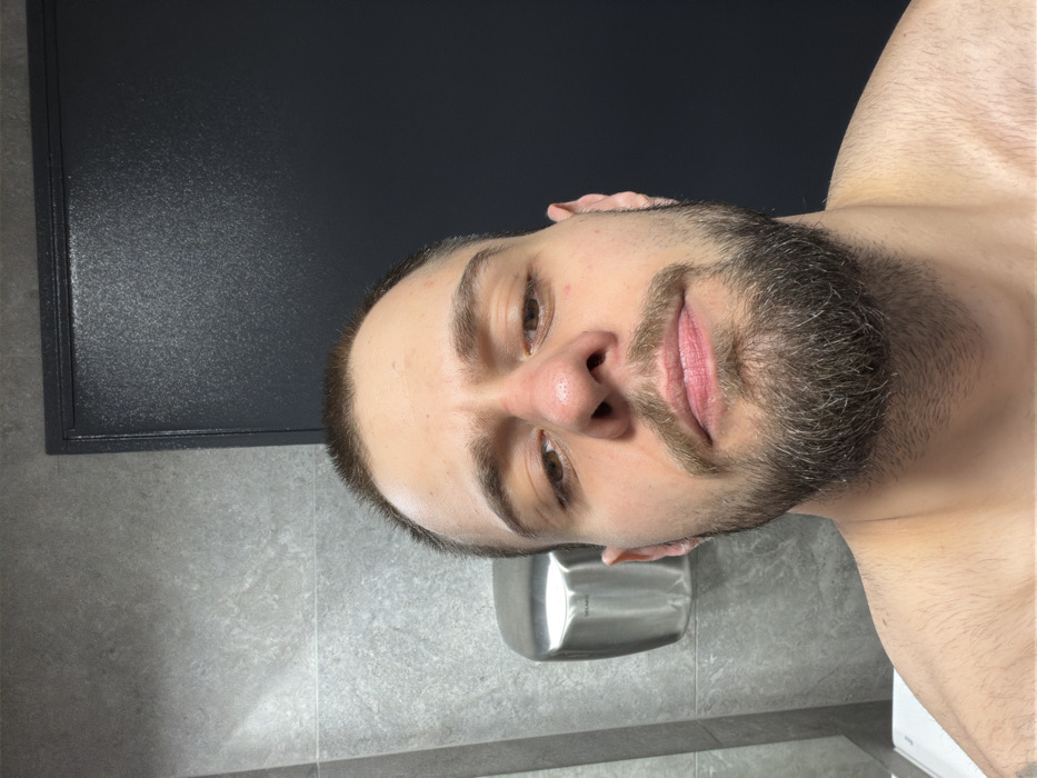 The image displays an adult male captured in a close-up, head-and-shoulders selfie within a public restroom in Plovdiv, Bulgaria. The subject has short, dark hair and a full, dark beard, presenting a neutral facial expression while looking directly into the camera. A subtle redness is noted on the bridge of his nose. He appears shirtless, with his bare upper chest and shoulders visible. The background on the left features gray tiled walls, against which a silver, wall-mounted DELABIE hand dryer is affixed. A dark gray, textured door with a distinct frame is visible on the right. A portion of a white sink basin or counter is discernible at the lower left edge of the frame.