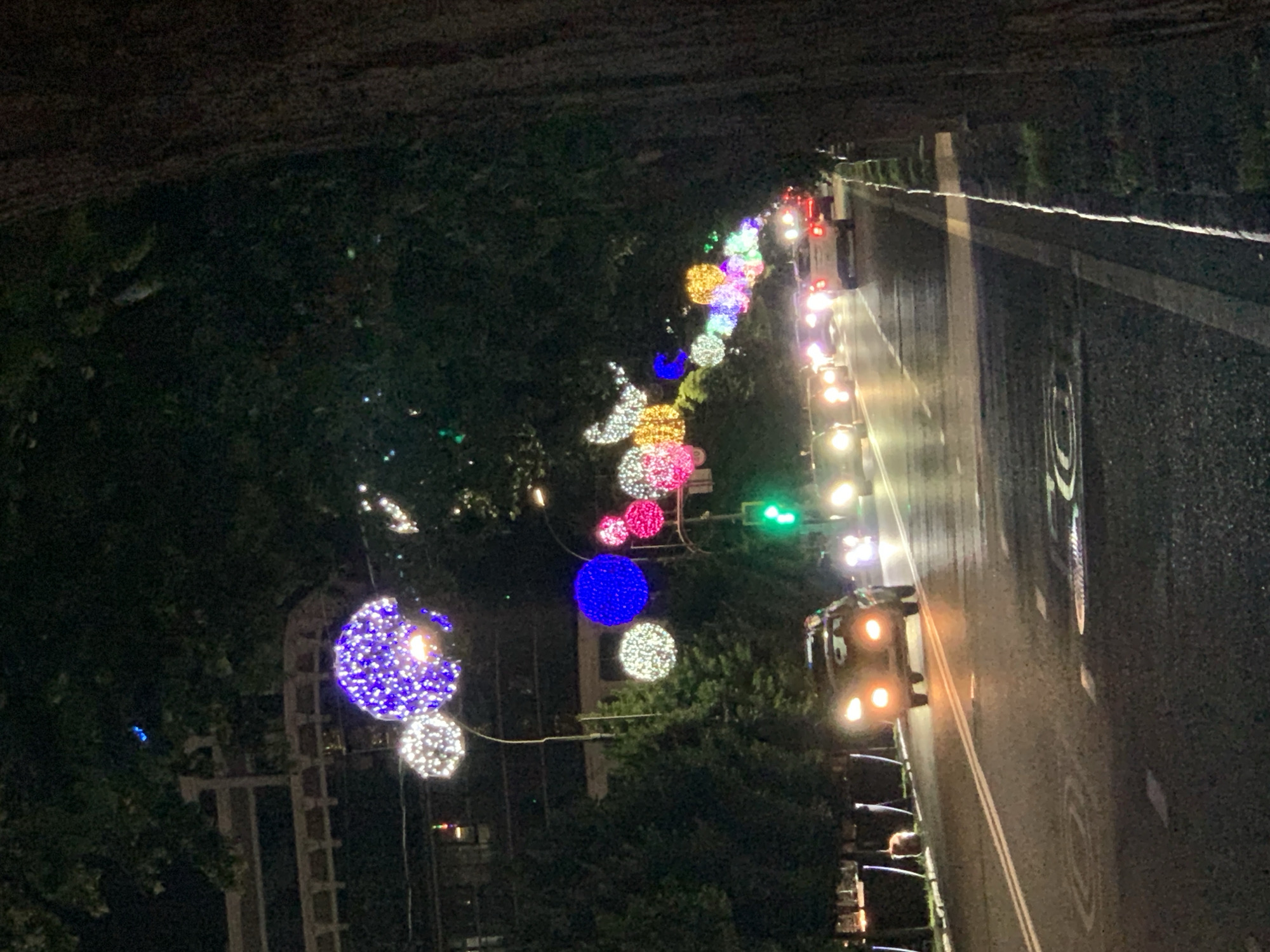 In the city of Unknown, Unknown, at night, a tree-lined street is decorated with illuminated spherical ornaments. The ornaments are suspended over the road, strung from what appear to be lampposts and tree branches. They are multi-colored, with prominent displays of blue, white, pink, and yellow lights. The street is wet, reflecting the lights of the vehicles and the decorative spheres. Several cars are visible on the road, their headlights and taillights illuminated. A traffic light, showing green, is visible in the background. The trees on either side of the road are dark and leafy, creating a canopy over the scene. The overall impression is a festive and somewhat atmospheric night scene on an urban street.
