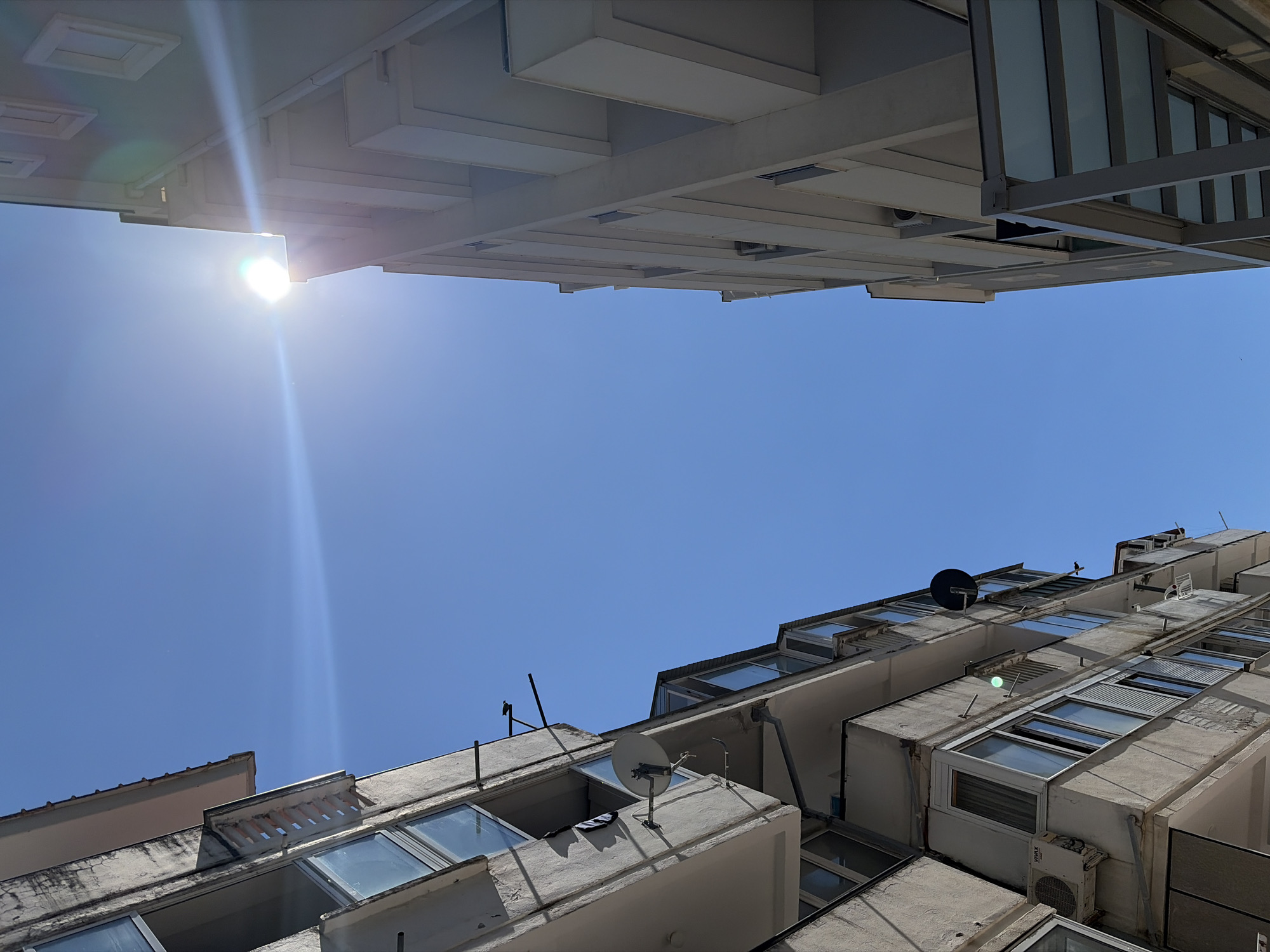 The image is an upward-angled shot of buildings in Budva, Montenegro, under a clear blue sky. The sun is visible at the top left, creating a lens flare. Two buildings are visible on the top and bottom of the view. The building at the bottom is more dilapidated, with multiple balconies, windows, satellite dish, and an air conditioning unit. The building at the top appears to be more modern and well-maintained, featuring gray metal construction and multiple square recesses.