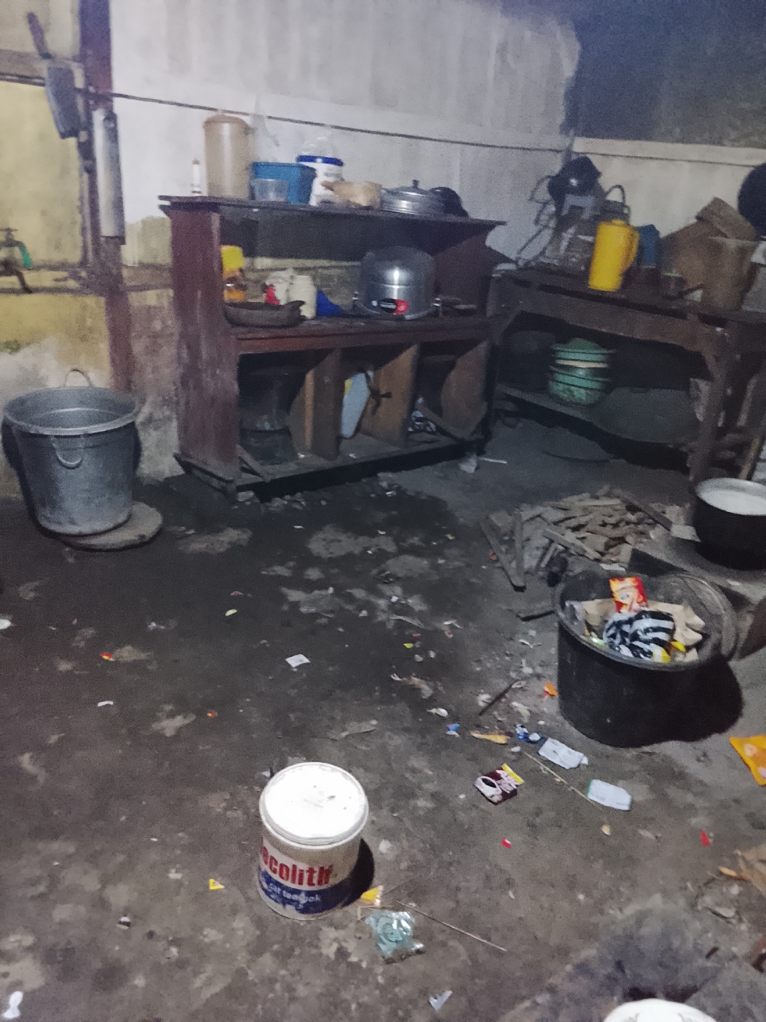 The image depicts a cluttered, dimly lit interior space that appears to be a kitchen or storage area. The floor is concrete and littered with debris, including paper, plastic wrappers, and what appears to be small pieces of wood. Several metal and plastic containers are visible, some containing what might be trash or other materials. In the foreground, a white bucket with red lettering is on the floor. In the mid-ground, a wooden shelf unit holds various items, including pots, containers, and what seems to be a yellow pitcher. To the right, a dark wooden table holds more containers and debris. A large grey metal bucket sits on the left side of the image, near a wall with a faucet. The walls are roughly plastered and uneven, with some staining. The overall impression is one of disarray and neglect. The location is identified as Mernek, Indonesia.