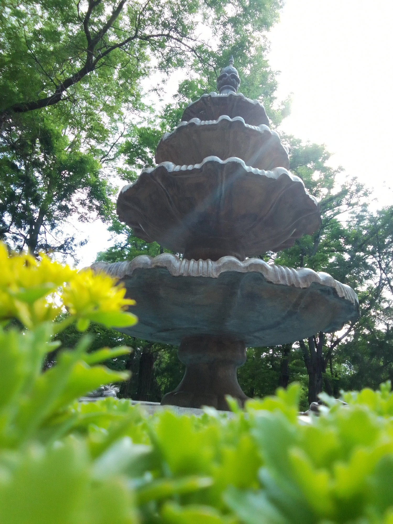 A tiered fountain made of bronze or stone stands in the center of the frame. The fountain has three levels, each with a scalloped edge. The top level has a decorative finial. Green foliage and yellow flowers are in the foreground, obscuring the base of the fountain and the ground. Trees with dense green leaves form the background, and the sky is bright and washed out with sunlight. The image appears to be taken at eye level, looking up at the fountain, with a shallow depth of field that blurs the foreground and background.