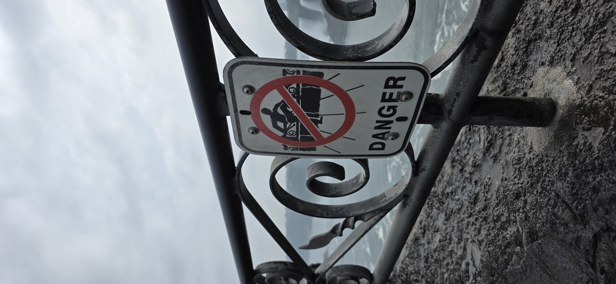 A vertical, eye-level, medium shot captures a weather-beaten "Danger" sign attached to a black metal railing. The sign, predominantly white with a red circular prohibition symbol, depicts a black silhouette of a person sitting on a bench, with a red line crossing out the image, indicating "no sitting on the bench." The word "DANGER" is printed in black capital letters below the symbol. The railing itself is decorative with ornate scrollwork, and it separates a wet, dark, textured ground surface from a body of water with visible waterfalls in the background. The sky above is overcast with soft gray clouds. The scene is located at the Niagara Falls in Canada, as indicated by the presence of the falls in the background and a typical tourist area railing and signage.