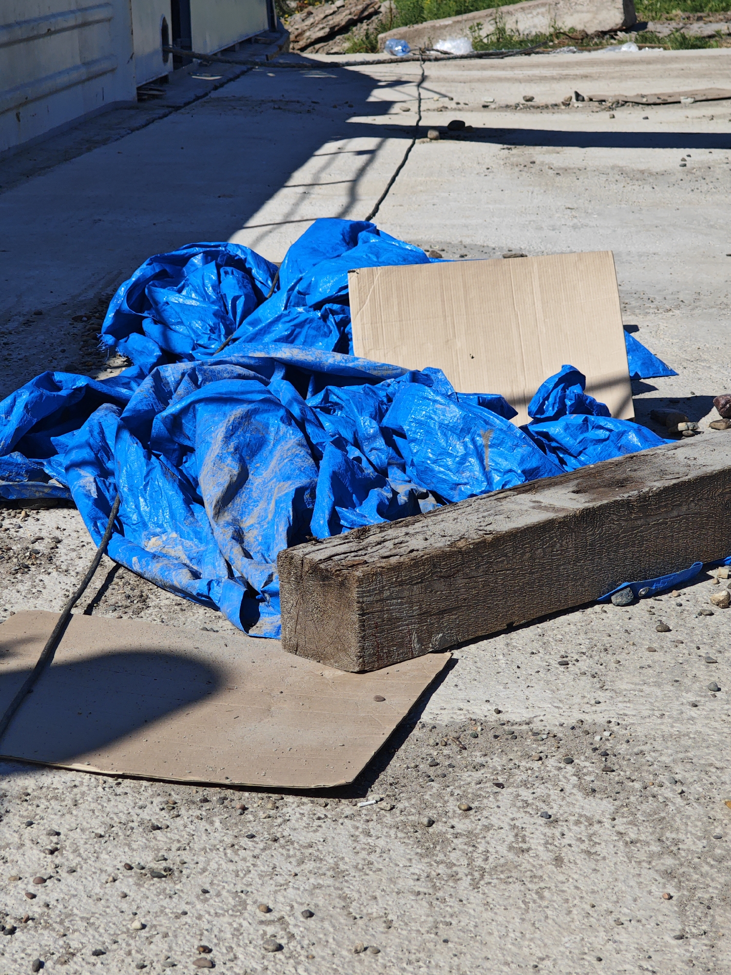 The image shows a pile of blue tarp covering what appears to be construction debris on a concrete surface. A weathered wooden beam rests on top of the tarp. A piece of cardboard is also present. The scene is outdoors, with shadows suggesting it is daytime. The background includes a light-colored building and some vegetation. The concrete surface shows signs of wear and tear, with scattered pebbles and dirt. There are no people visible in the image.