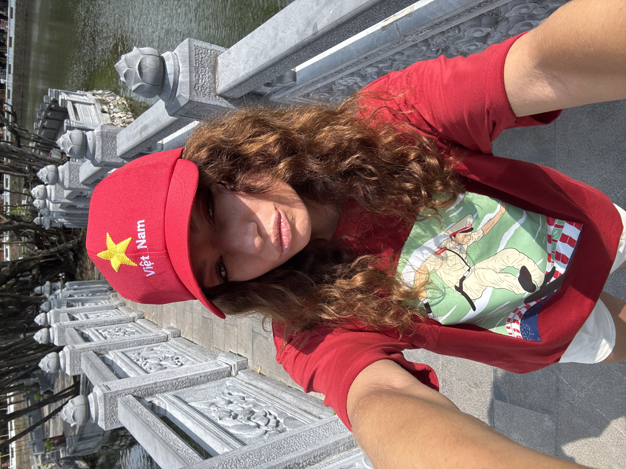 A woman takes a selfie outdoors. She wears a red baseball cap embroidered with a gold star and the words "Việt Nam." She also wears a red t-shirt with a graphic print. She stands on a stone bridge, likely the Huc Bridge (Cầu Thê Húc), as it appears to have lotus-shaped stone pillars and carved panels. Visible in the background is the Hoàn Kiếm Lake (Hồ Hoàn Kiếm) in Hà Nội, Vietnam.
