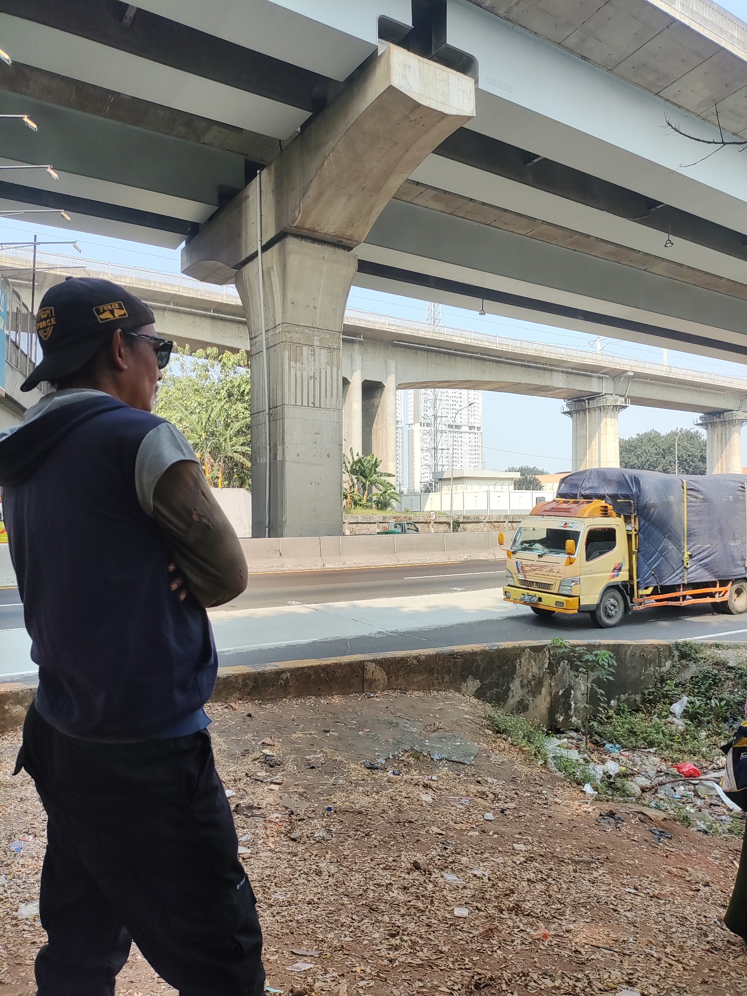 A man wearing a navy blue hooded jacket over a grey shirt and black pants stands with his arms crossed, facing away from the camera. He also wears a black baseball cap with patches and sunglasses. In the background, a yellow truck with a blue tarp covering its cargo is driving on a highway under an elevated structure. Buildings and trees are visible in the distance. The foreground is a dirt path littered with debris, including leaves and plastic waste. The scene appears to be in an urban or semi-urban area, possibly near a construction site or an underpass.