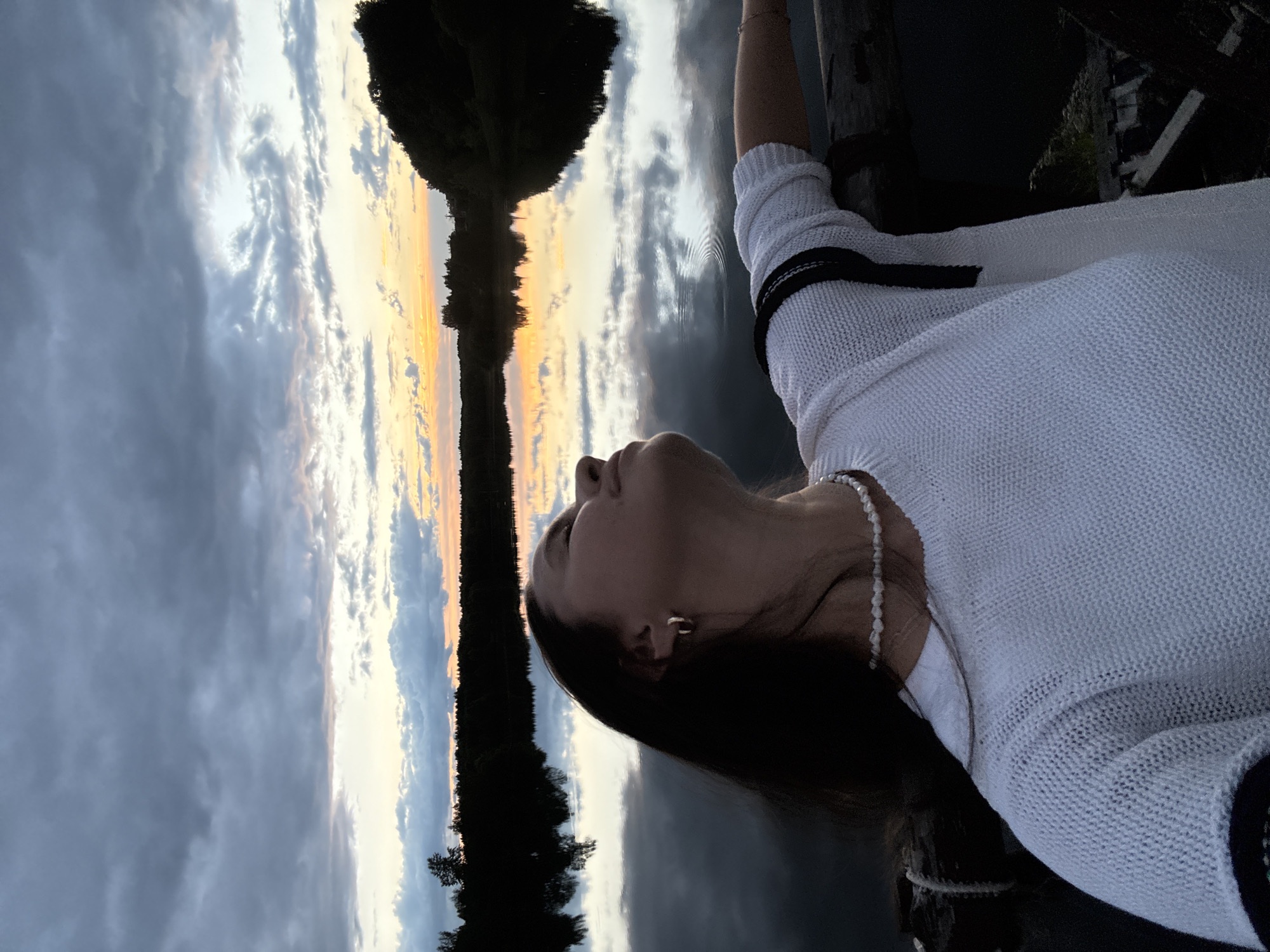 This vertical photograph captures a contemplative moment against a stunning natural backdrop, likely during sunrise or sunset, in Kamysh, Russia.
The main subject is a woman, positioned on the right side of the frame, shown in profile. She is looking upwards and slightly towards the left. Her face is partially shadowed, emphasizing her dark, long hair which falls behind her. She wears a textured, white knitted sweater with black trim on the cuff of her visible arm, and a delicate pearl necklace is visible around her neck. A small, simple hoop earring is also seen in her ear.
The background is dominated by a dramatic sky and its reflection in a calm body of water, presumably a lake or river. The horizon line runs horizontally across the middle of the image, dividing the sky above from its mirrored image in the water below. The sky transitions from warm golden and orange tones on the left, indicative of the sun's position, to cooler shades of grey and blue on the right. Various cloud formations are scattered across the sky, some wispy, others more substantial.
A dark, silhouetted landmass, likely a distant shoreline with trees, stretches across the far left of the image, also perfectly reflected in the still water. The water's surface shows subtle ripples in places, distorting the reflections slightly but adding to the dynamic visual.
The overall impression is one of peaceful observation, with the woman seemingly lost in thought or appreciation of the natural beauty of the changing sky. The time of day is clearly either early morning or late evening due to the vivid and transitional colors of the light. No visible text is present in the image.