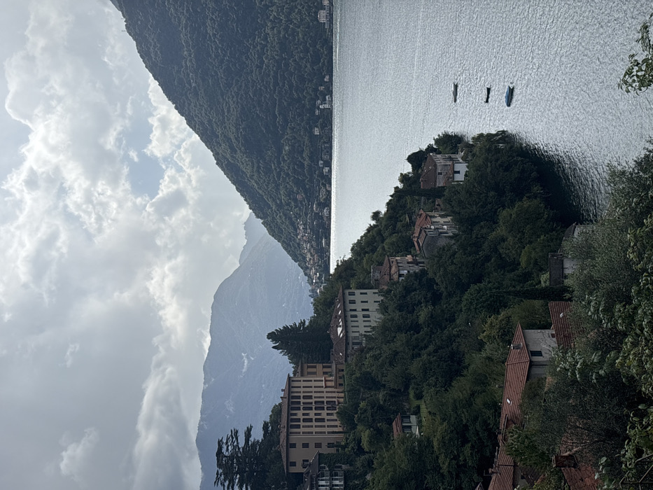 The image displays an elevated view of Lake Como within the city of Nesso, Italy, on an overcast day. The upper portion of the frame is dominated by a sky with extensive light-gray cloud cover, interspersed with areas of brighter sky towards the right. Multiple layers of mountains, predominantly dark green with dense tree cover, frame the scene; a prominent, steeply sloped mountain occupies the right side, descending directly into the lake, while additional hazy mountain ranges recede into the distance on the left and center.
The central and lower-right sections feature a large body of water, Lake Como, exhibiting ripples across its surface and reflecting a silvery-gray light. Three small, unoccupied boats are visible on the water in the middle-right: the top boat is light-colored, the middle boat is dark, and the bottom boat is blue and covered.
The left and central foreground are characterized by a steep, tree-covered incline forming the shoreline, dense with various green deciduous and coniferous trees. Numerous buildings of varying sizes are nestled within this verdant landscape. A prominent, light-colored (beige or cream) multi-story building with multiple window rows stands on an elevated terrace in the mid-left. Below and to its right, another lighter-colored multi-story building with a red-tiled roof is partially visible. Further down the slope towards the lake's edge, several smaller residential structures with terracotta-tiled roofs are interspersed among the trees. On the distant opposite shore, a continuous line of buildings is discernible, extending into the valley between the mountains. No individuals, discernible actions, or interactions are present in the image.