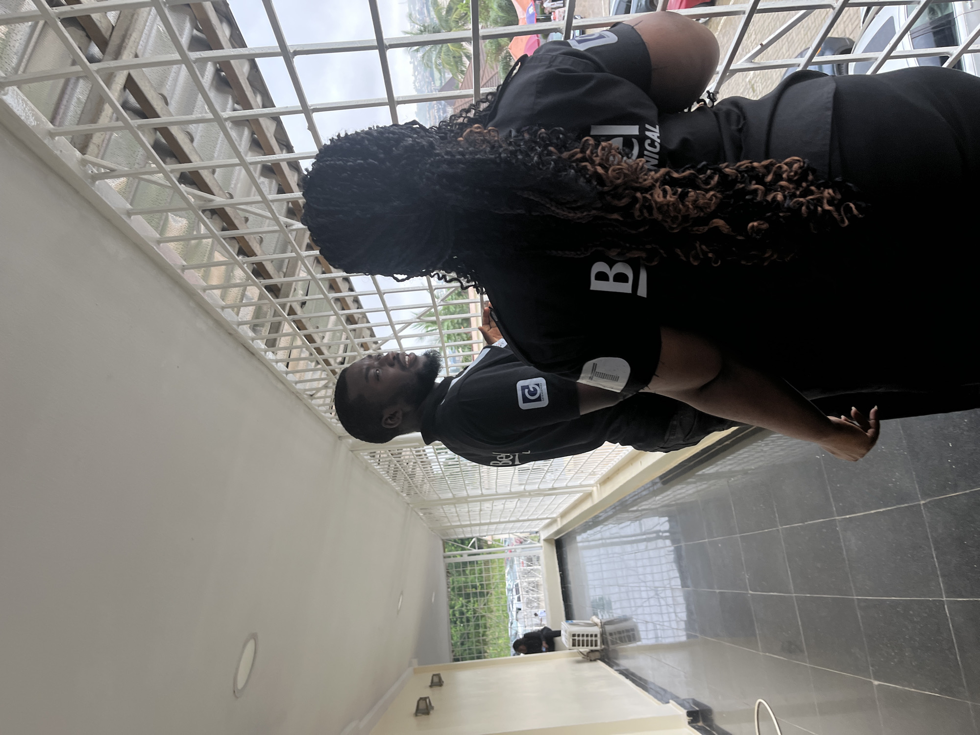 Two individuals, a man and a woman, are standing on a covered walkway, facing away from the camera. They are both wearing black shirts with a logo on the left sleeve and lettering on the back. The man has short dark hair and a beard, and he is looking to his right. The woman has long, dark, braided hair with some lighter brown highlights, and she is looking to her left. They appear to be engaged in a conversation. The walkway has a dark tiled floor and a white ceiling with fluorescent lights. A white air conditioning unit is visible on the left side of the walkway. The walkway is bordered by a white metal grate that offers a view of parked cars, trees, and buildings in the distance, suggesting an urban or suburban environment in Abuja, Nigeria. The sky is overcast.